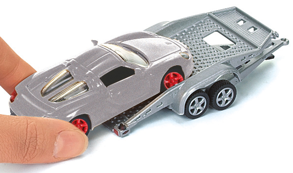 Ein Spielzeugauto auf einem grauen Anhänger, gehalten von einer Hand. Das Auto ist silberfarben mit roten Rädern.