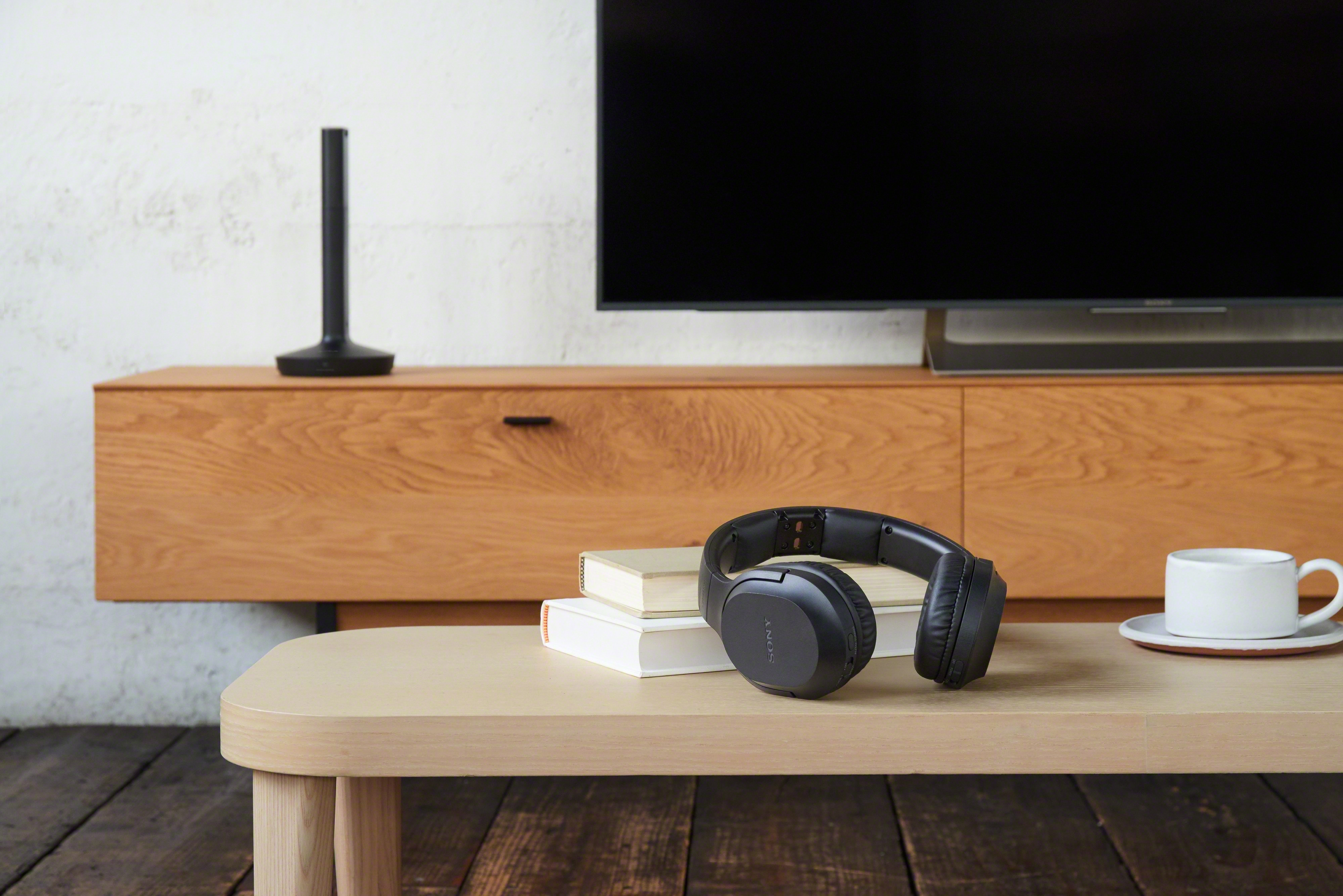 Casque sur une table, avec une TV et un meuble en arrière-plan. Livres et tasse à proximité.