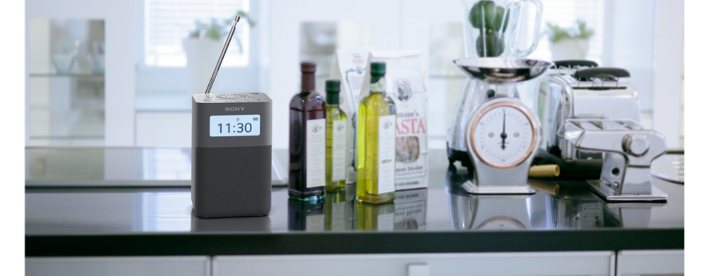 Une radio noire Sony affiche 11h30 sur un comptoir de cuisine, avec des bouteilles d'huile et des aliments en arrière-plan.