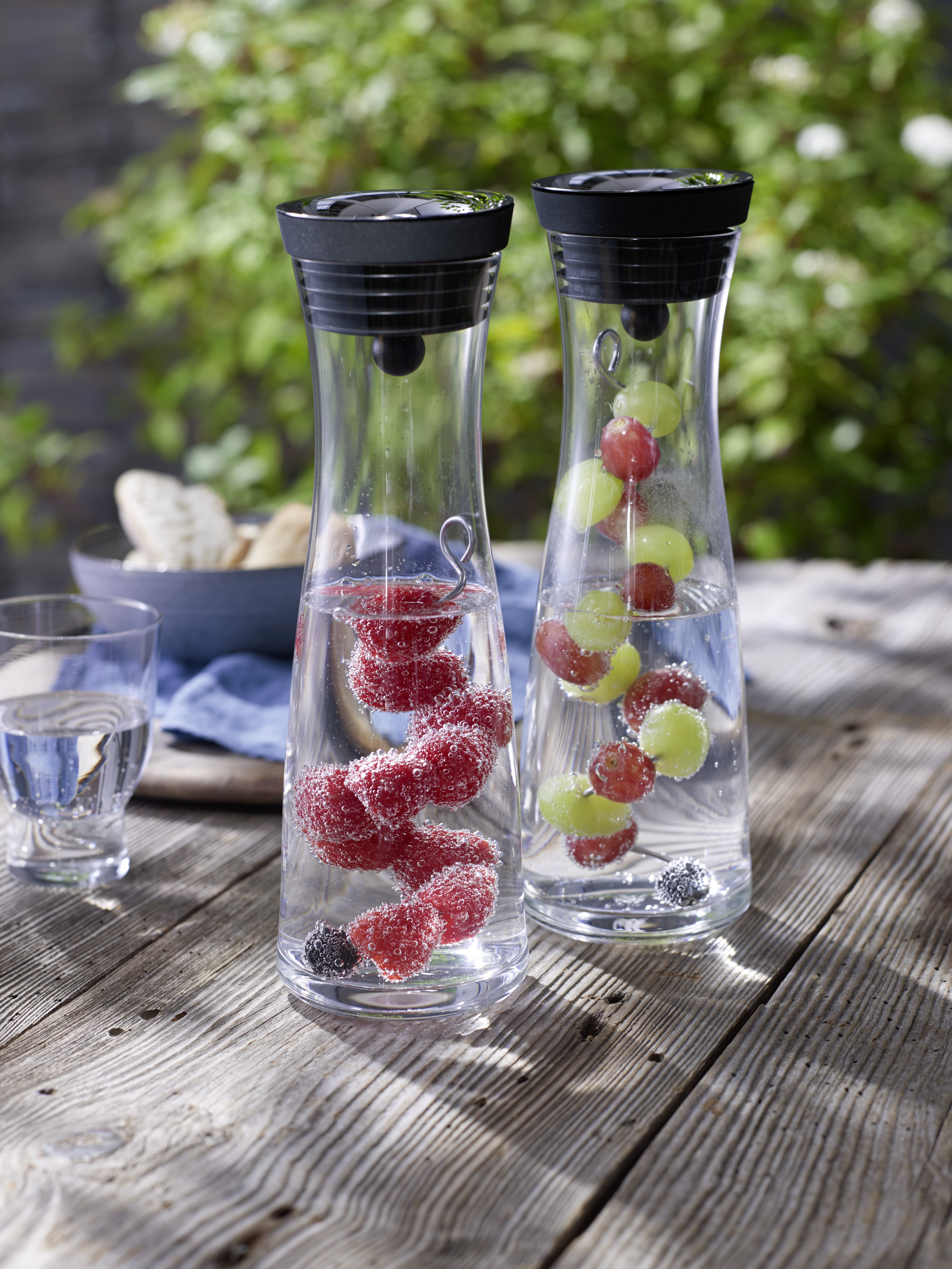 Zwei Glaskaraffen mit Früchten stehen draußen auf einem Holztisch. Ein Glas Wasser daneben.