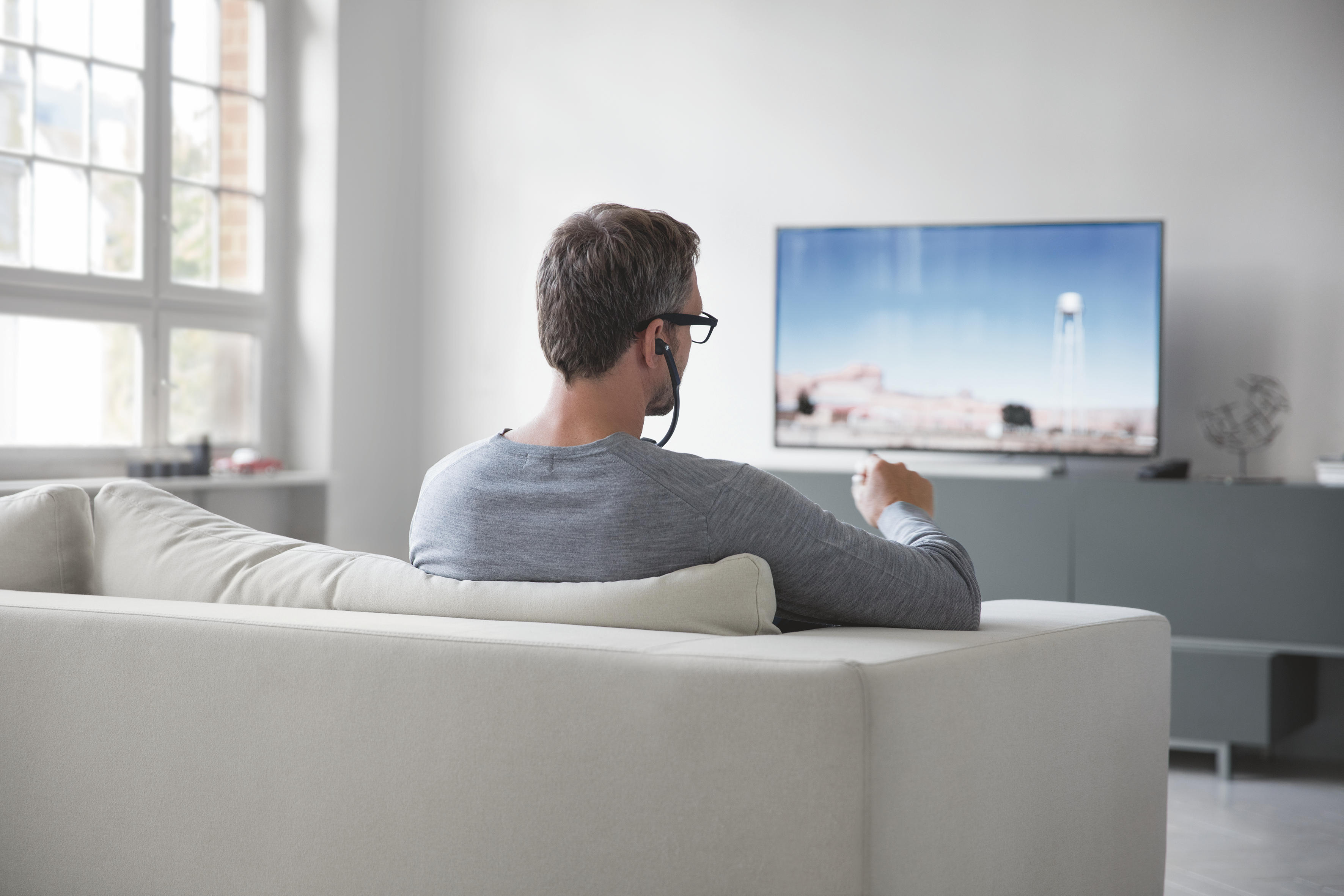 Ein Mann sitzt auf einem Sofa und trägt Kopfhörer. Er schaut fern. Der Fernseher zeigt eine hellblaue Szene.