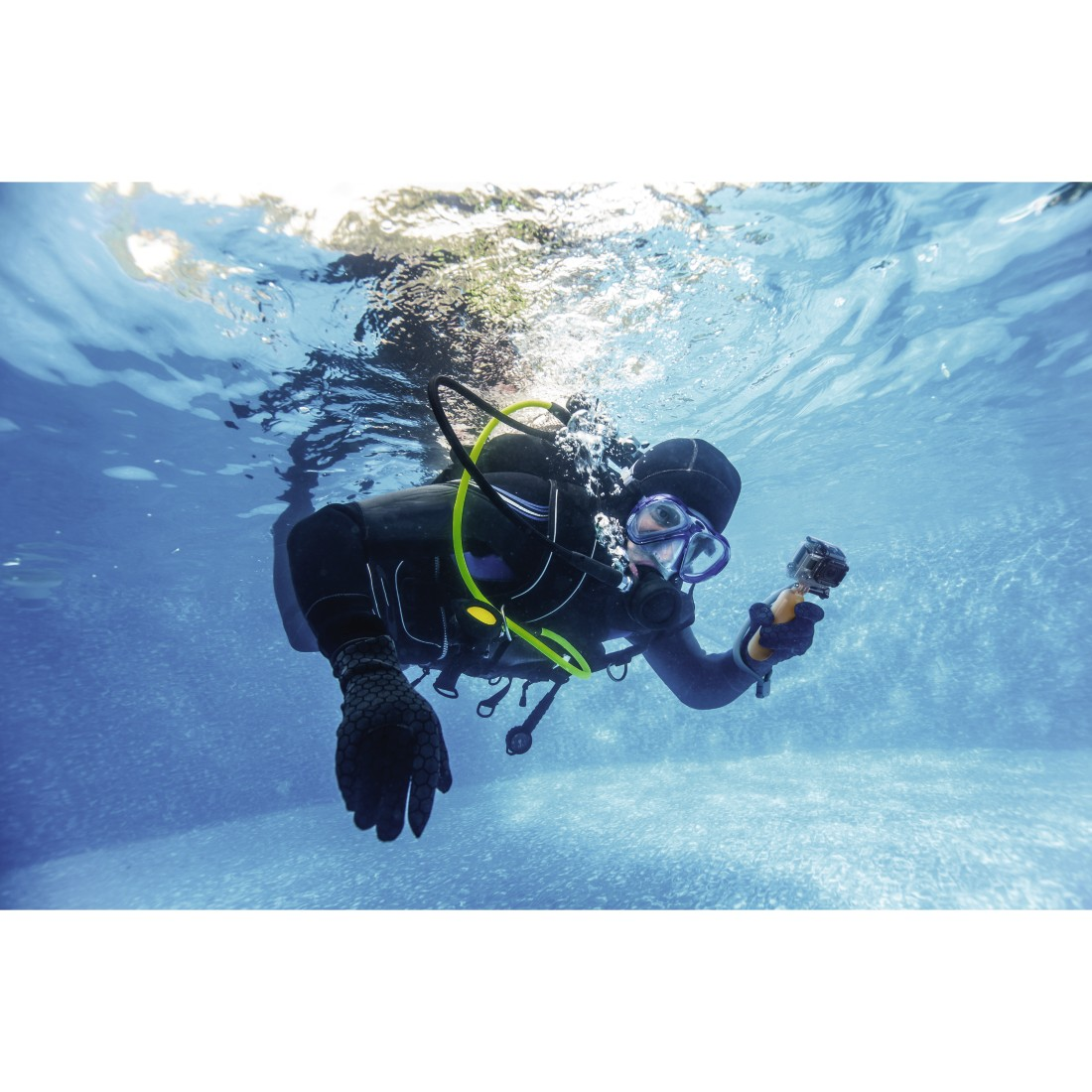 Scuba diver in full gear underwater, holding a camera. Bright blue water and sunlight above.