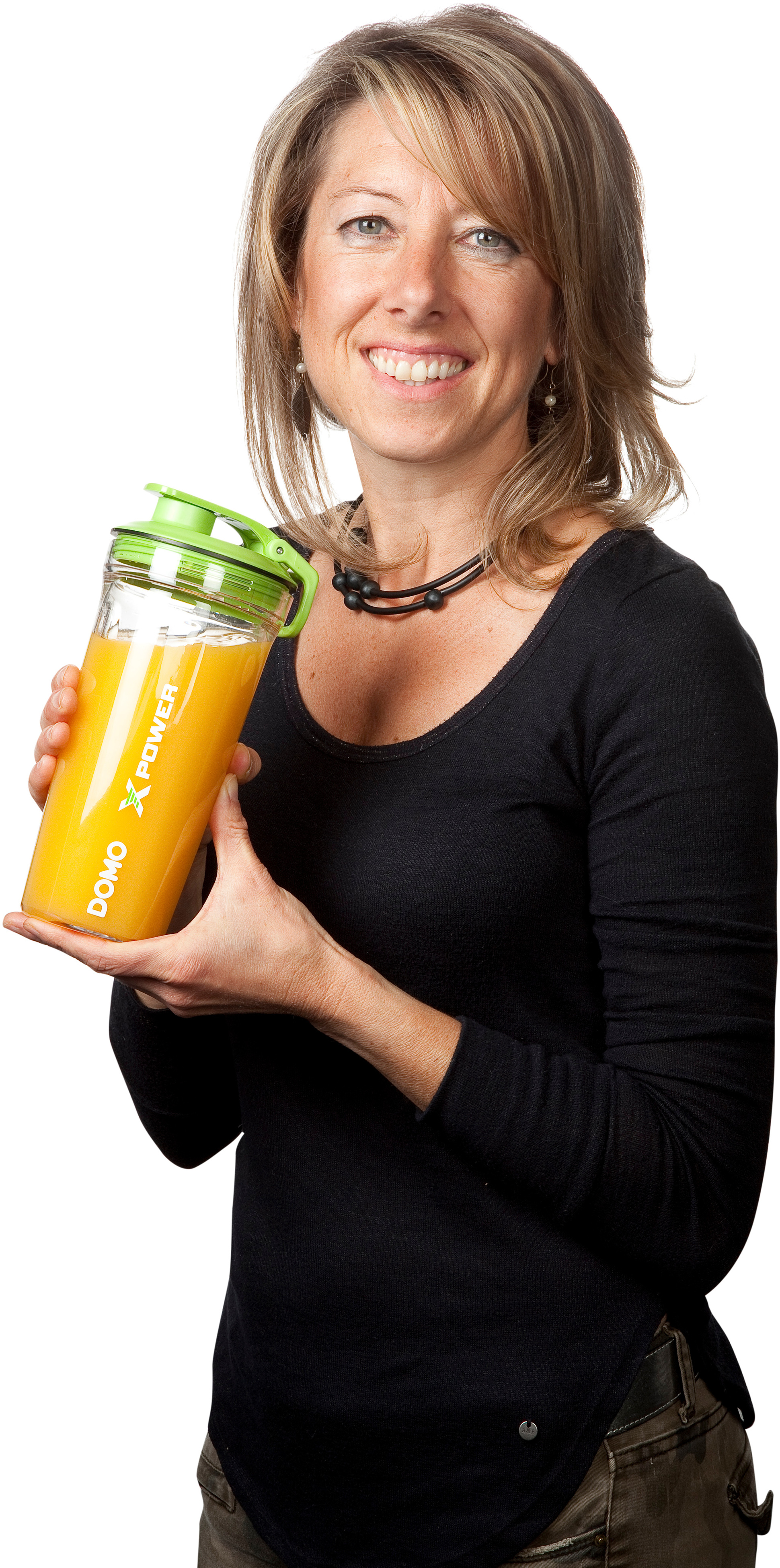 Une femme tient un shaker de jus avec du liquide orange. Elle sourit, portant une chemise noire et un pantalon foncé.