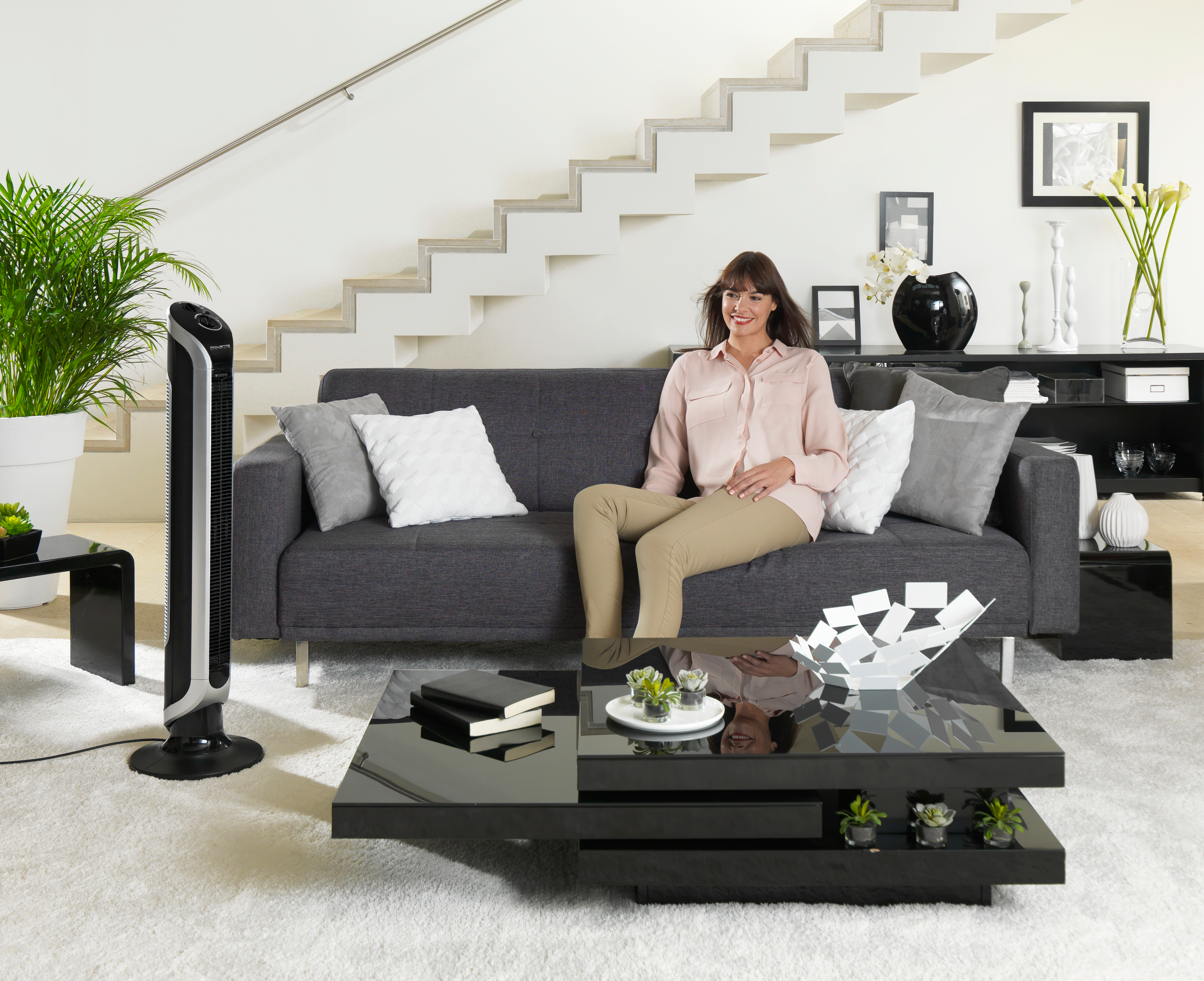 Une femme assise sur un canapé gris. Un ventilateur à gauche, une table basse devant et un escalier.