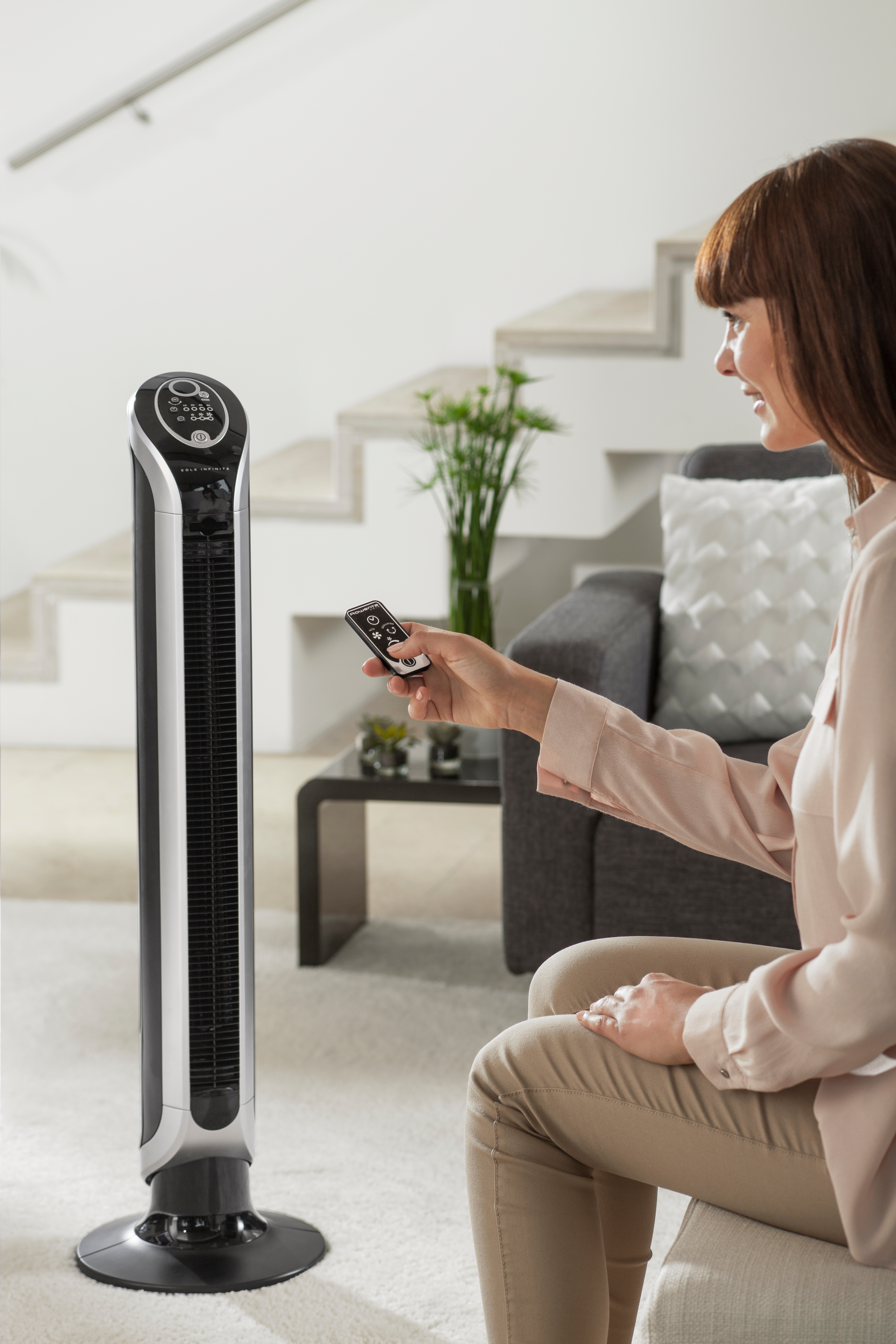 Une femme utilise une télécommande pour un ventilateur haut, argenté et noir.
