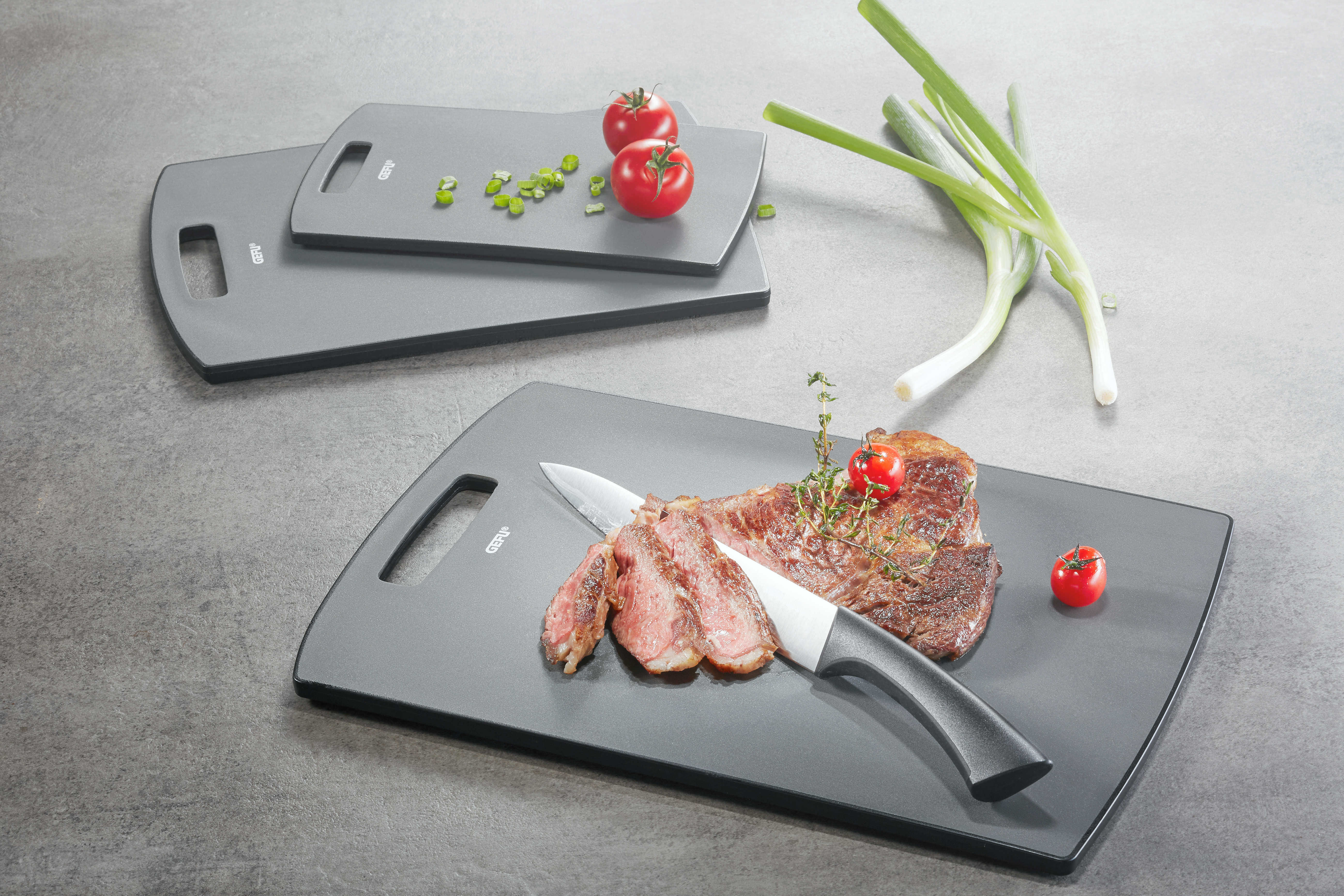 Graue Schneidebretter mit Steak, Messer und Tomaten. Zwei kleinere Bretter sind sichtbar.