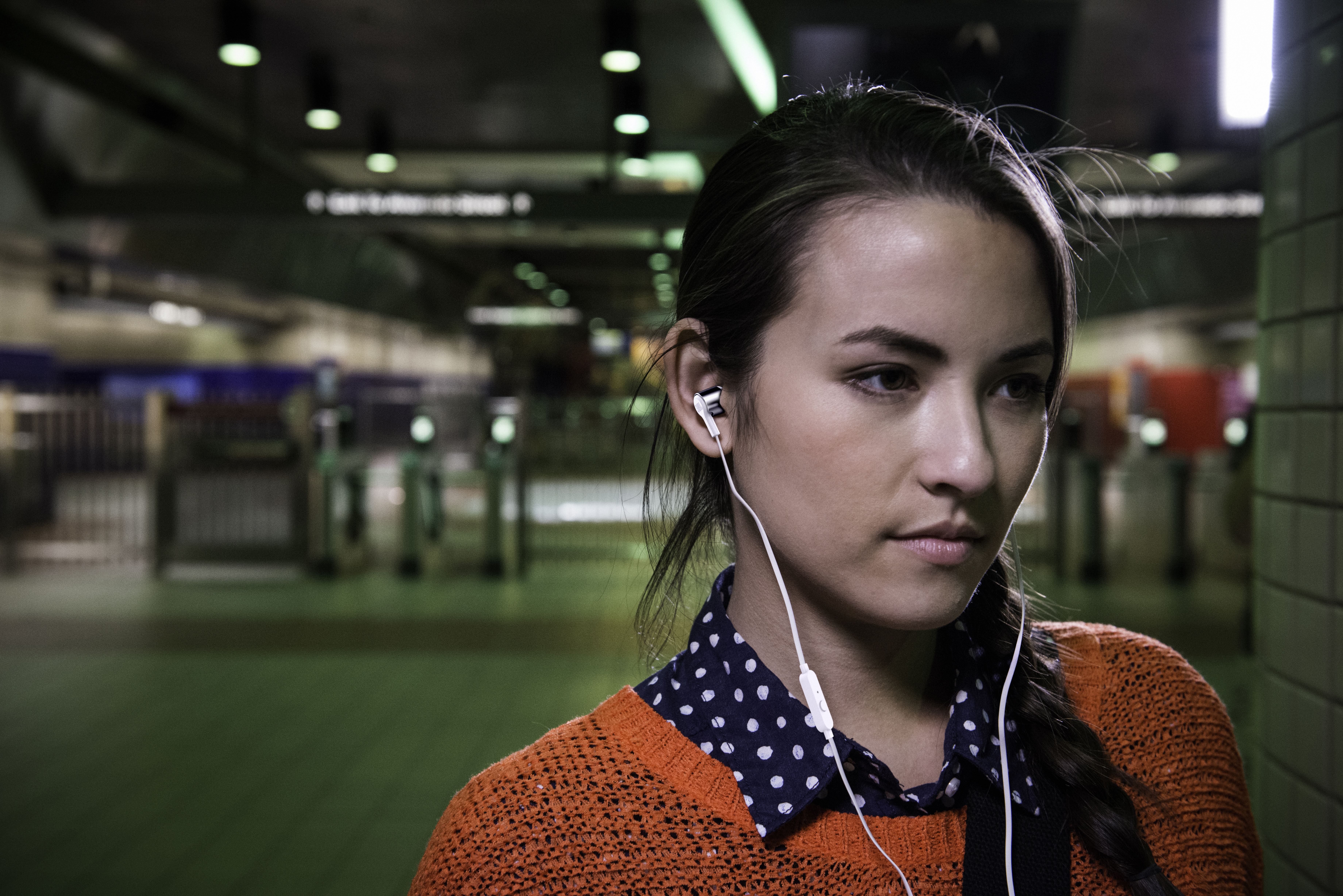 Eine Frau in einem orangefarbenen Pullover mit Kopfhörern in einer U-Bahn-Station, die konzentriert wirkt.