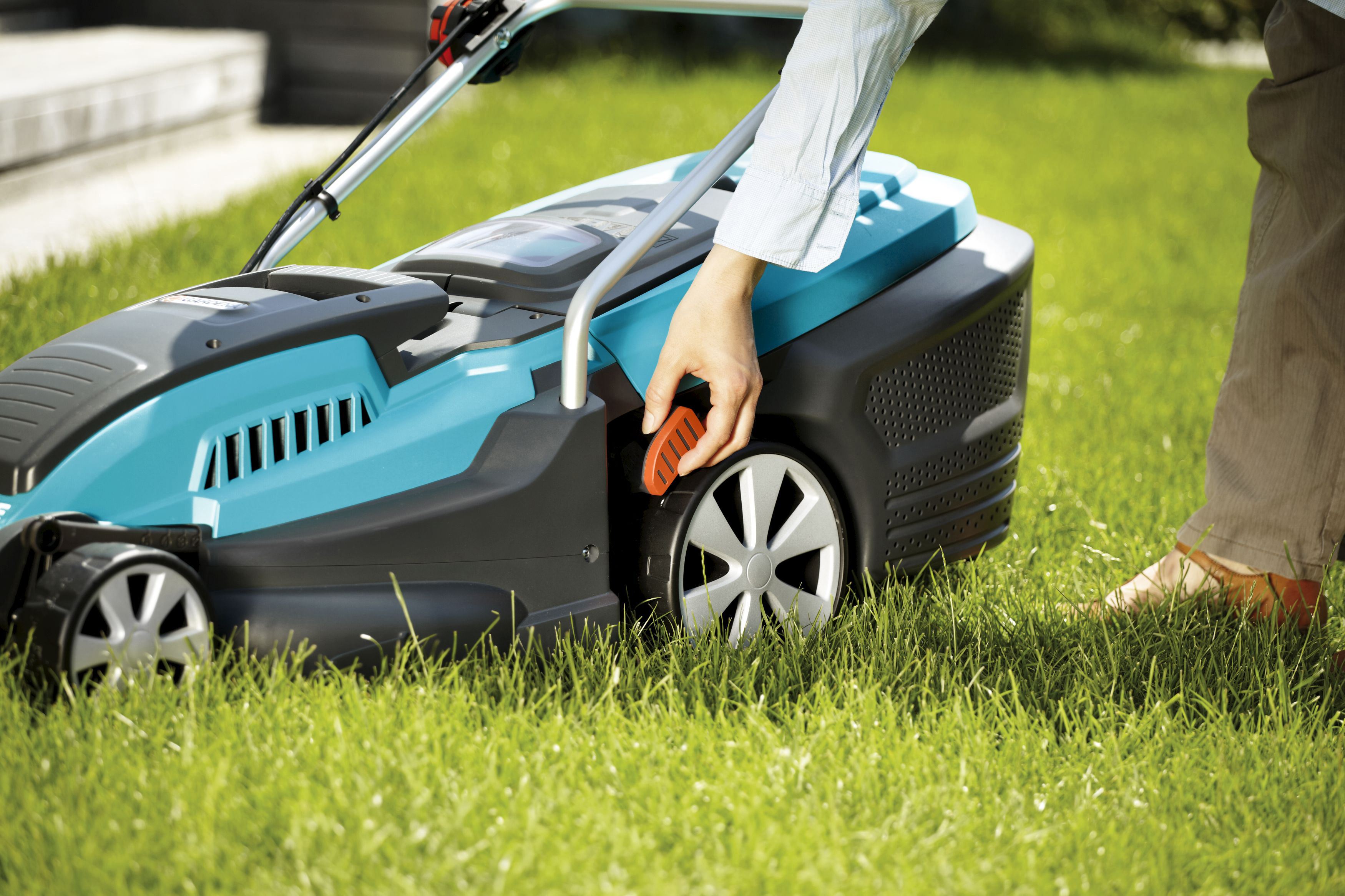 Person benutzt einen teal Rasenmäher auf grünem Gras. Oranger Hebel an der Seite.