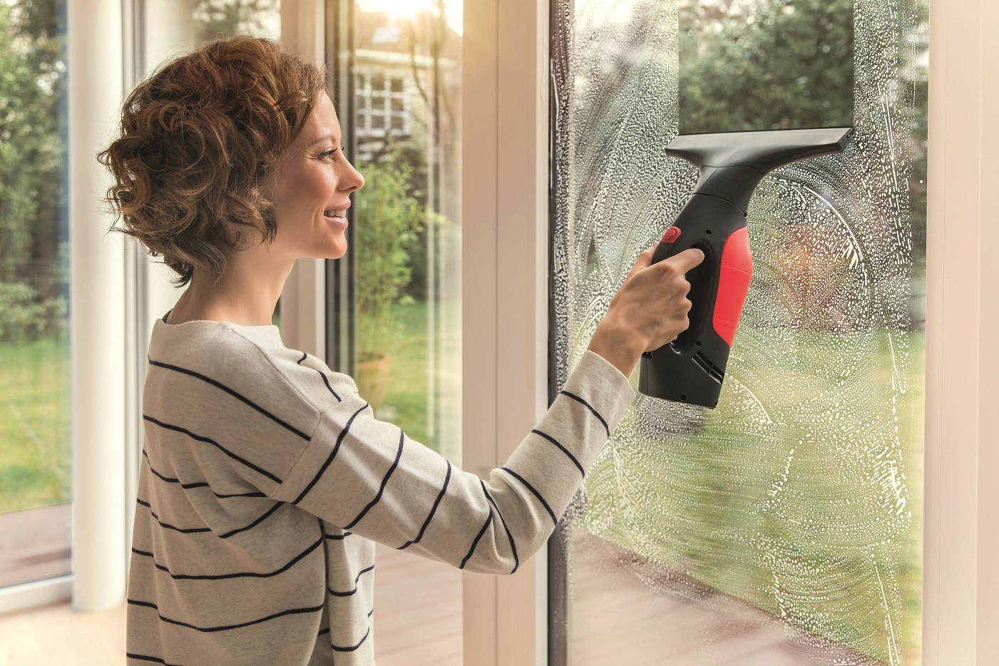 Frau putzt Fenster mit Abzieher. Draußen ist ein verschwommener grüner Garten zu sehen.