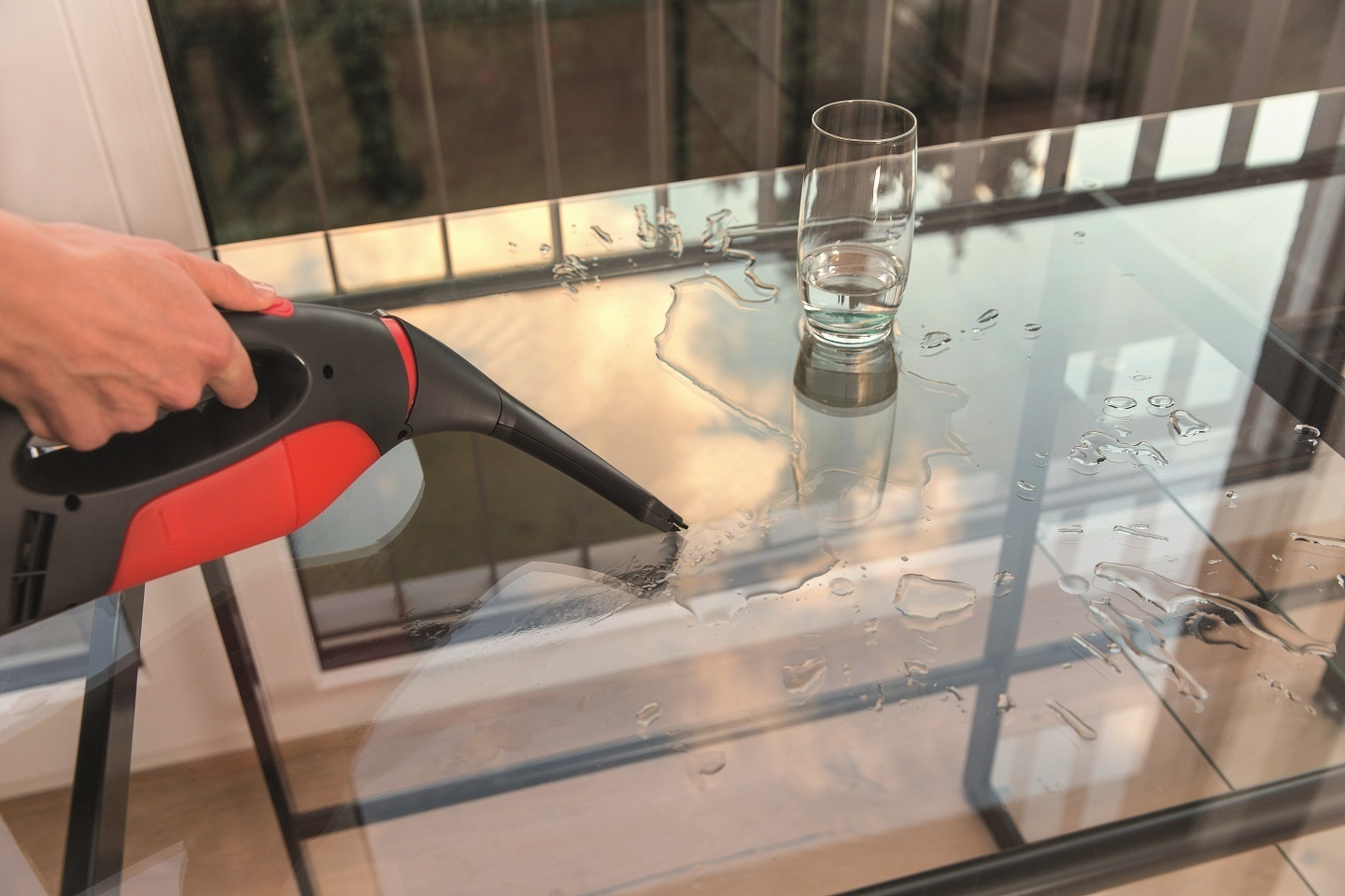 Frau reinigt Glastisch. Rotes und schwarzes Reinigungswerkzeug. Glastisch mit Wasserflecken. Fenster im Hintergrund.