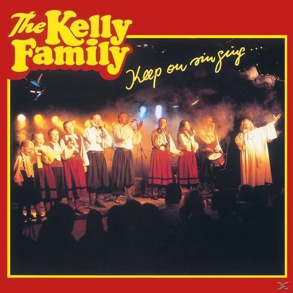 Albumcover: The Kelly Family singt auf der Bühne, Bandmitglieder in traditioneller Kleidung.