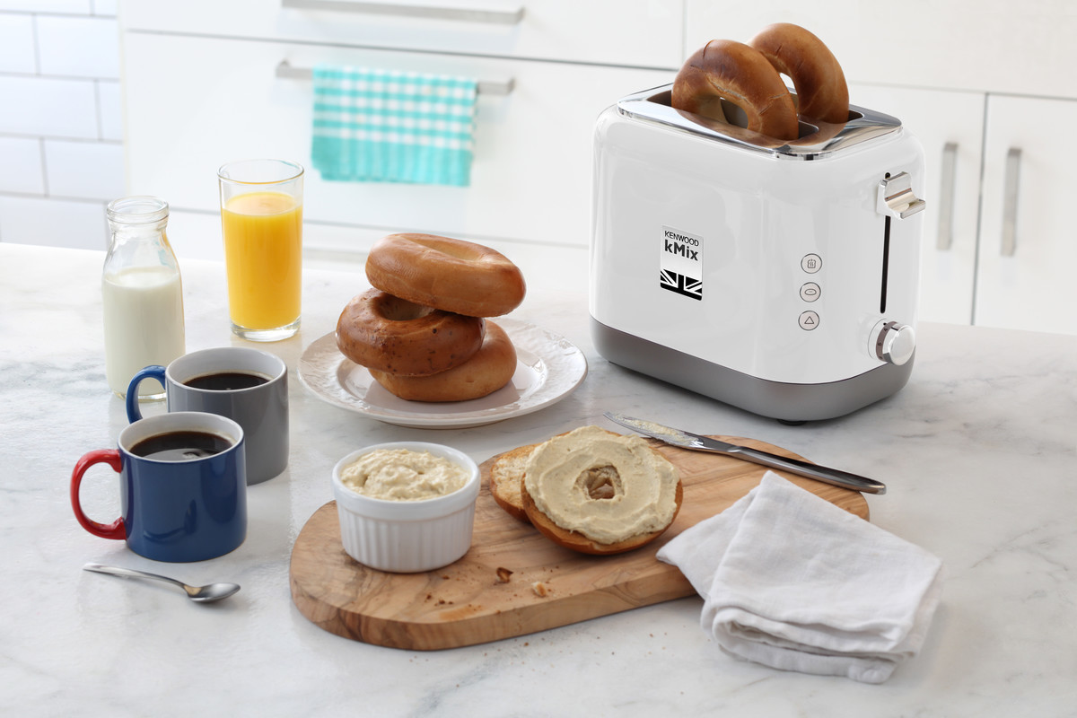 Scène de petit-déjeuner : Bagels, toasts, beurre, café, jus d'orange, lait et un grille-pain blanc sur un comptoir.