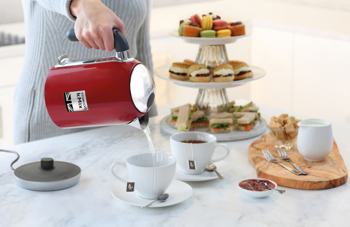 Frau gießt Wasser aus einem roten Wasserkocher in eine Teetasse. Sandwiches und Kuchen auf einem Tisch.