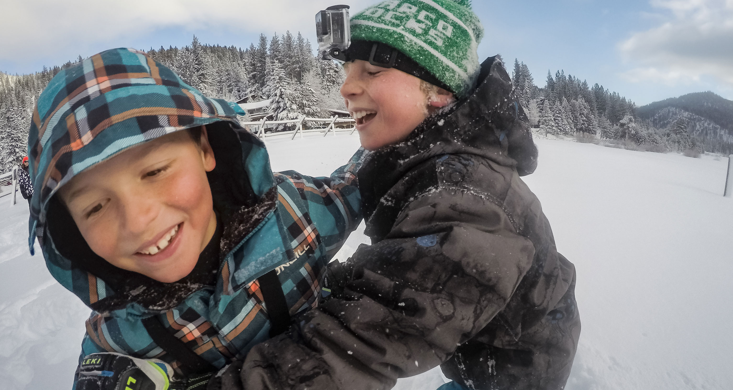 Két fiú nevet, hóban játszanak. Egyik zöld sapkát és GoPro-t visel. Havas háttér, fák.