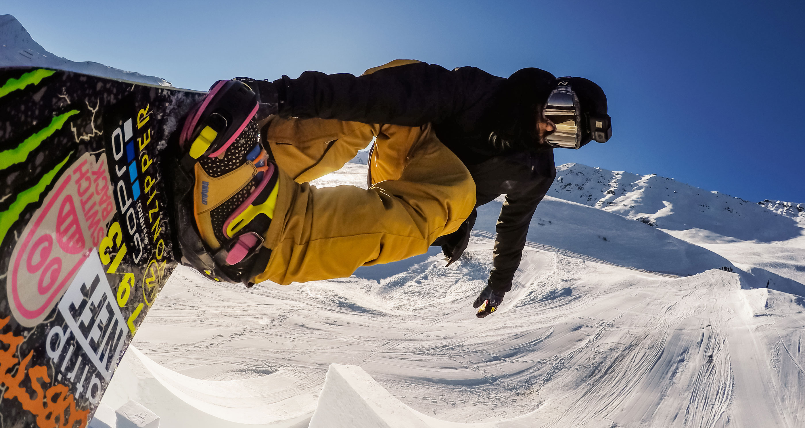 Snowboardos a levegőben, havas tájon. Égszínkék ég. A deszkán matricák vannak, a lovas fekete kabátot és szemüveget visel.