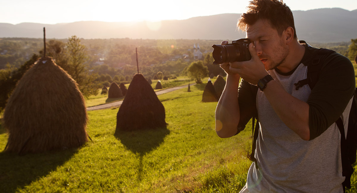 Egy férfi fényképet készít a tájról szénaboglyákkal és egy faluval a távolban, naplementekor.
