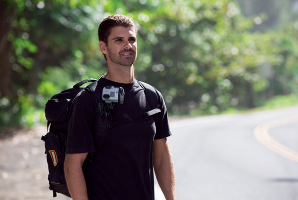 A man in a black shirt walks on a road, wearing a backpack and a camera on his chest.