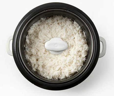 Rice cooker containing cooked white rice. The lid is a white, round shape in the middle of the rice.