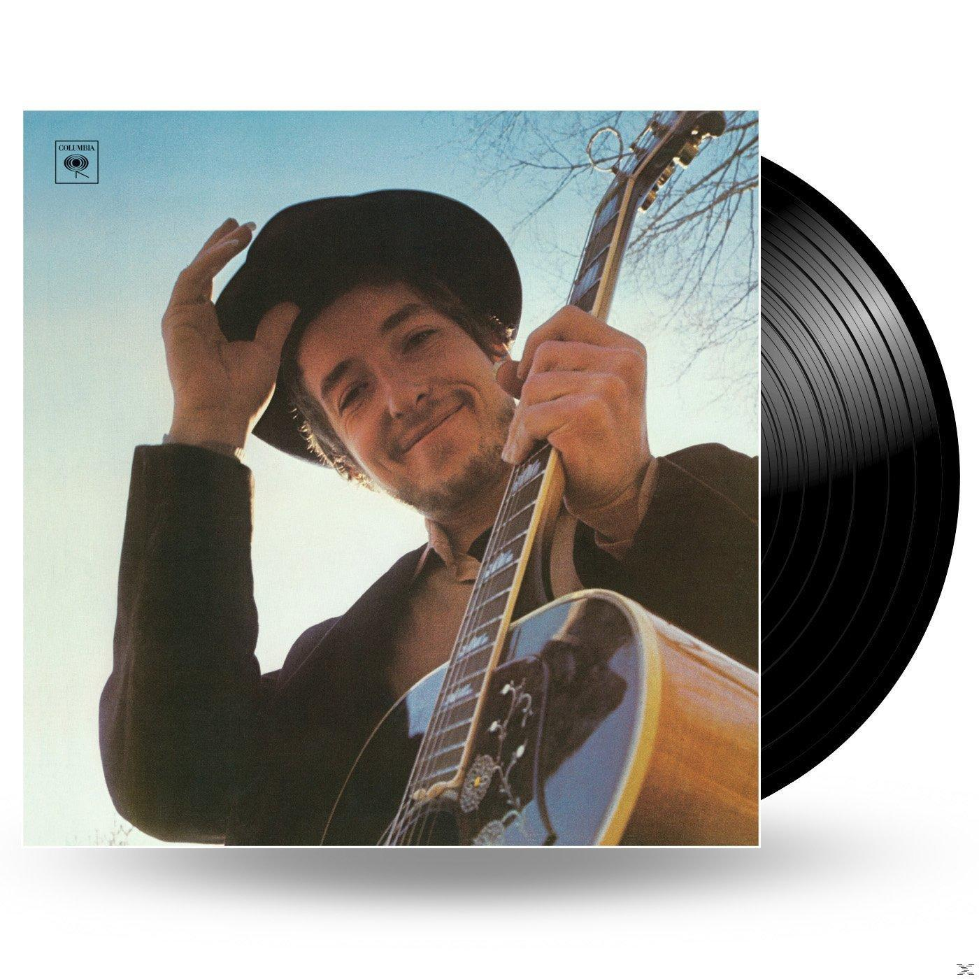 Albumcover mit Bob Dylan, der eine Gitarre hält und lächelt. Eine Schallplatte daneben.