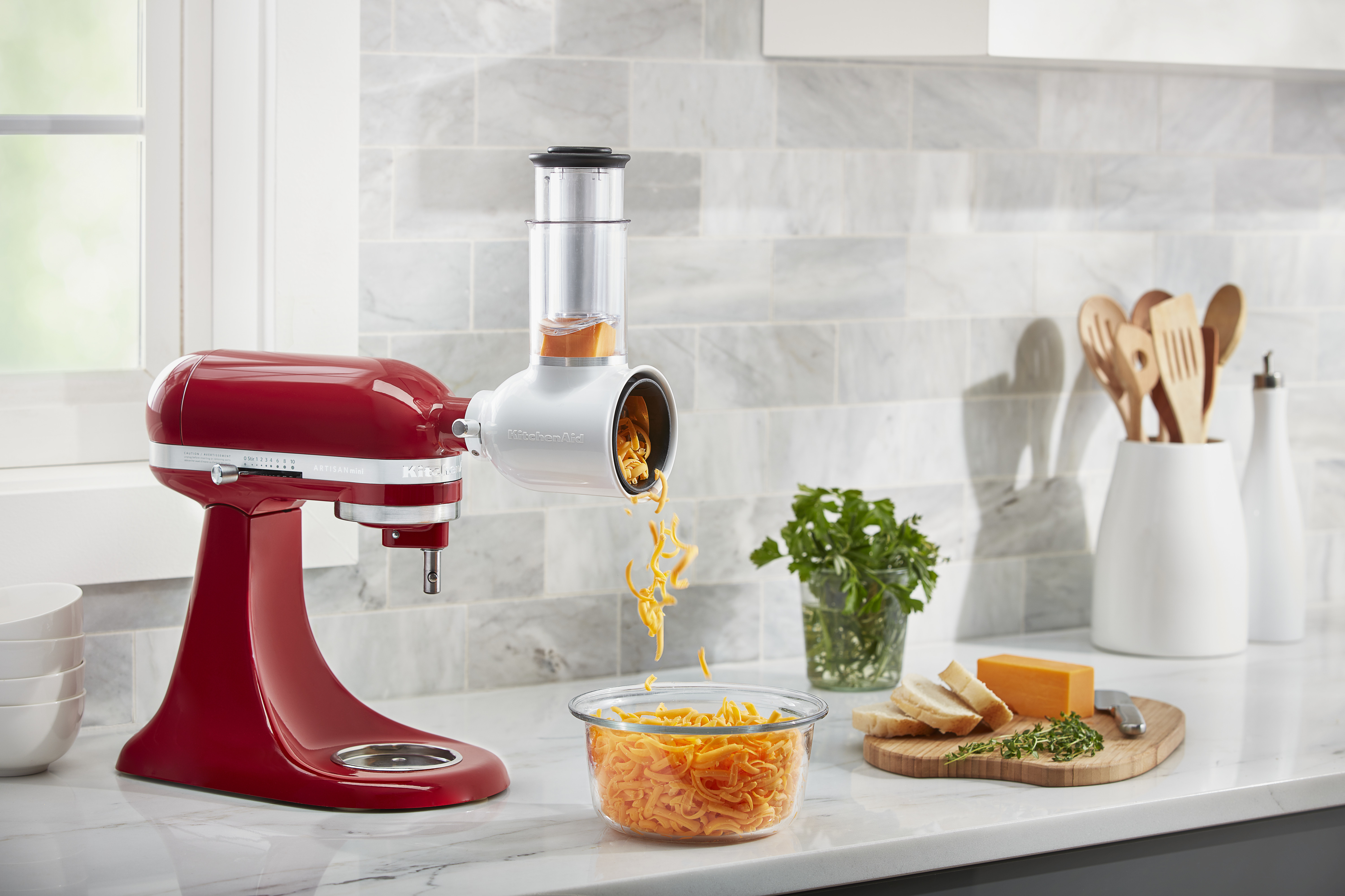 Ein roter KitchenAid-Mixer mit Reibeaufsatz, Käse und Gemüse auf einer Arbeitsplatte.