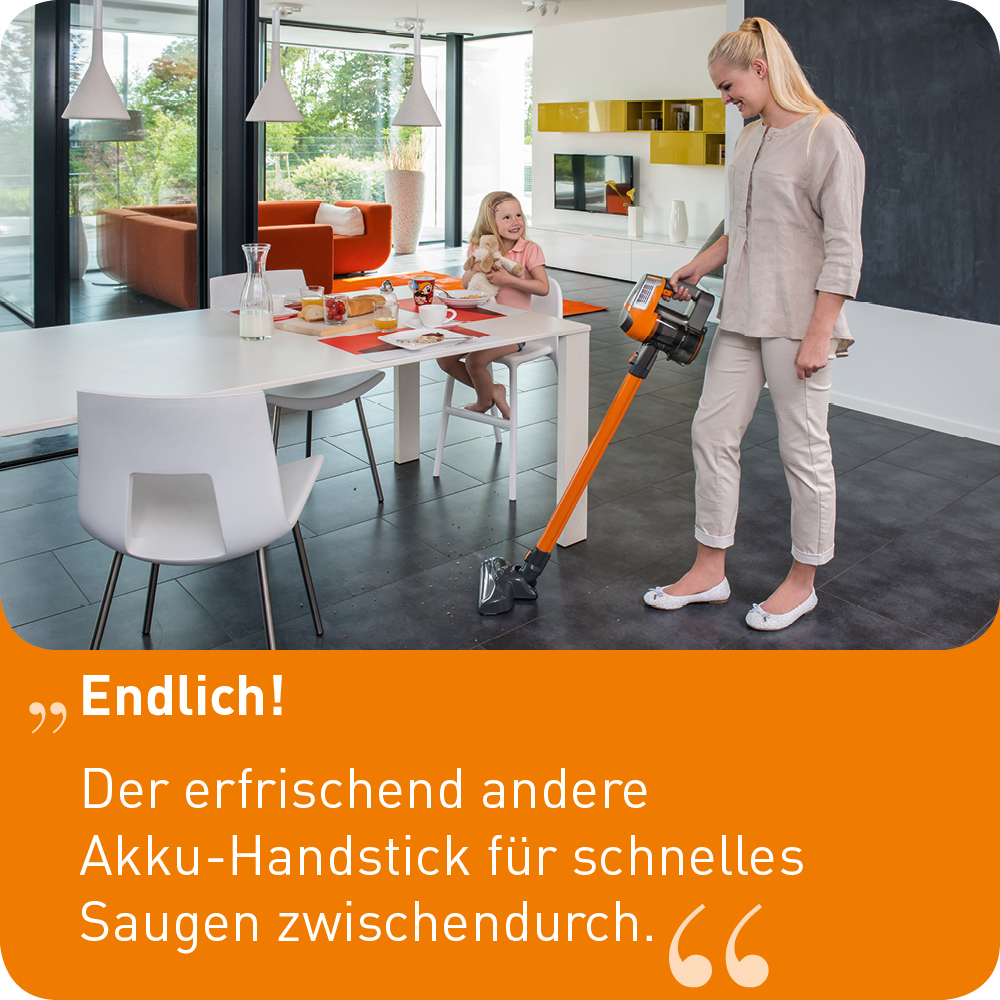 Frau staubsaugt, Kind isst am Tisch. Oranger Staubsauger im Vordergrund.
