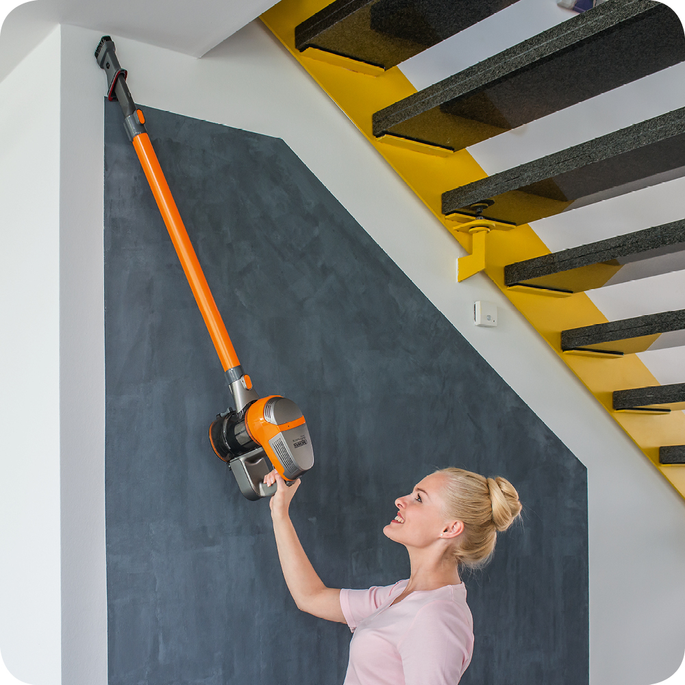 Eine Frau benutzt einen orangefarbenen Staubsauger an einer grauen Wand.