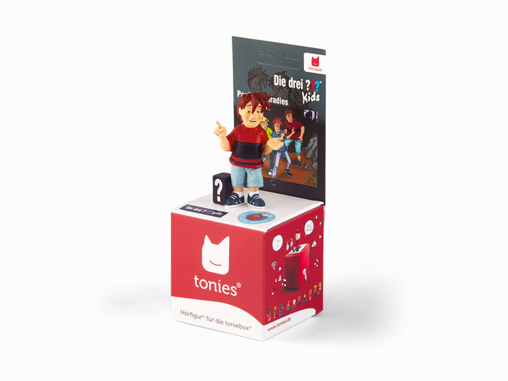 Eine Tonies-Figur eines Jungen in einem roten Hemd steht auf einer roten Box. Die Box hat das Tonies-Logo und ein Bild eines roten Audiogeräts.