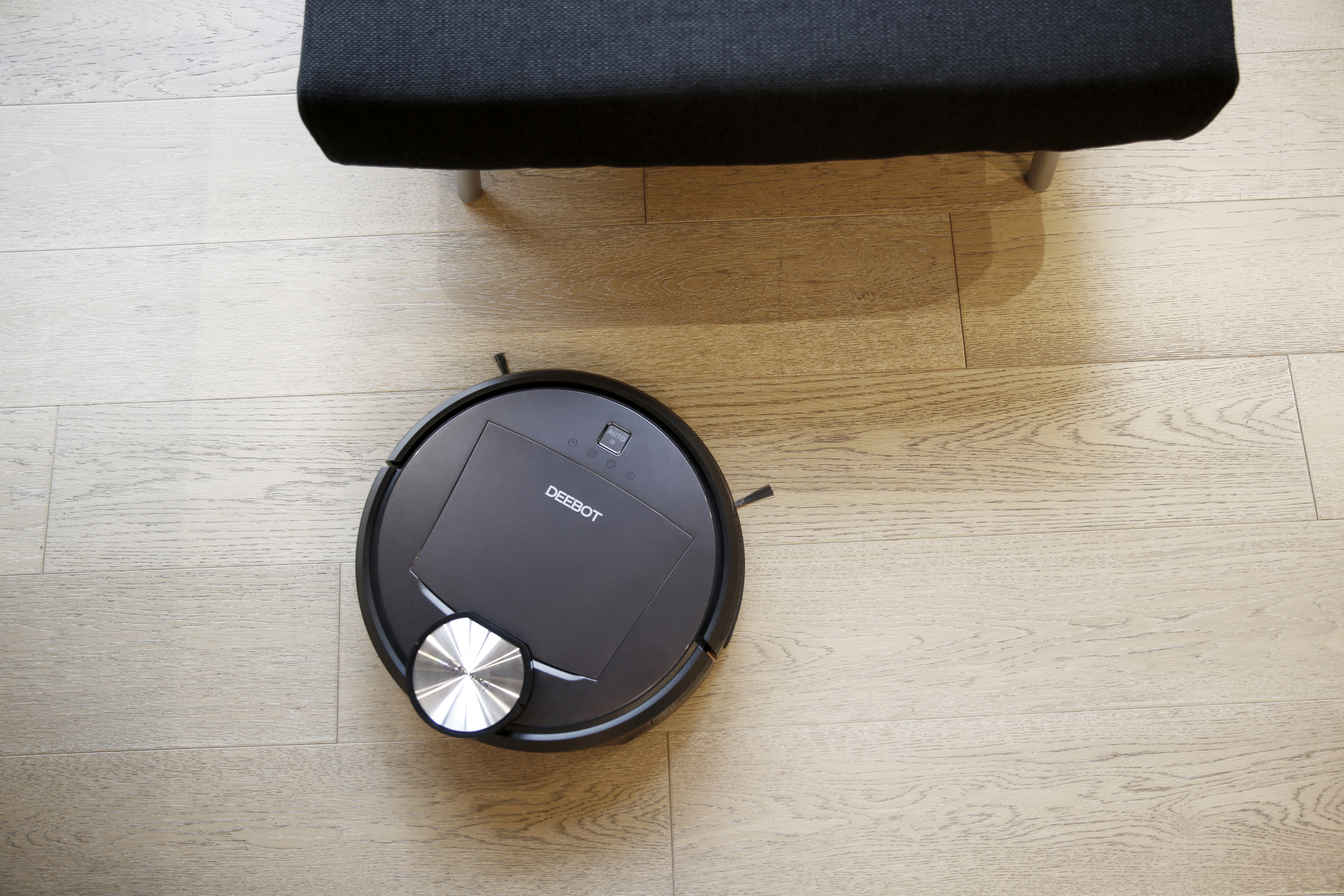 Schwarzer DEEBOT Roboter-Staubsauger auf Holzboden, in der Nähe eines schwarzen Stuhls.