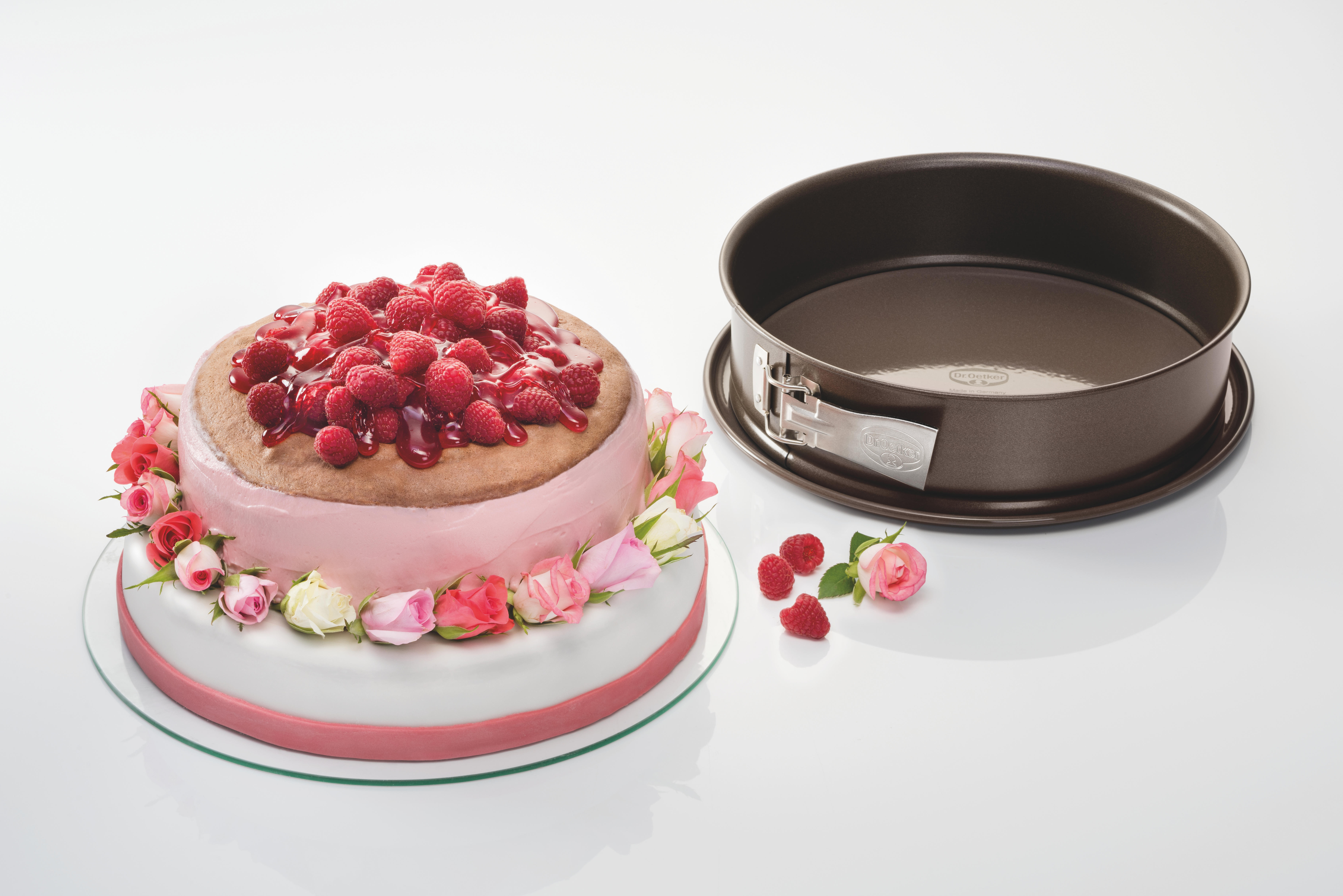 Kuchen mit rosa Glasur und Himbeeren, neben einer Kuchenform. Rosen sind auf dem Teller. Weißer Hintergrund.