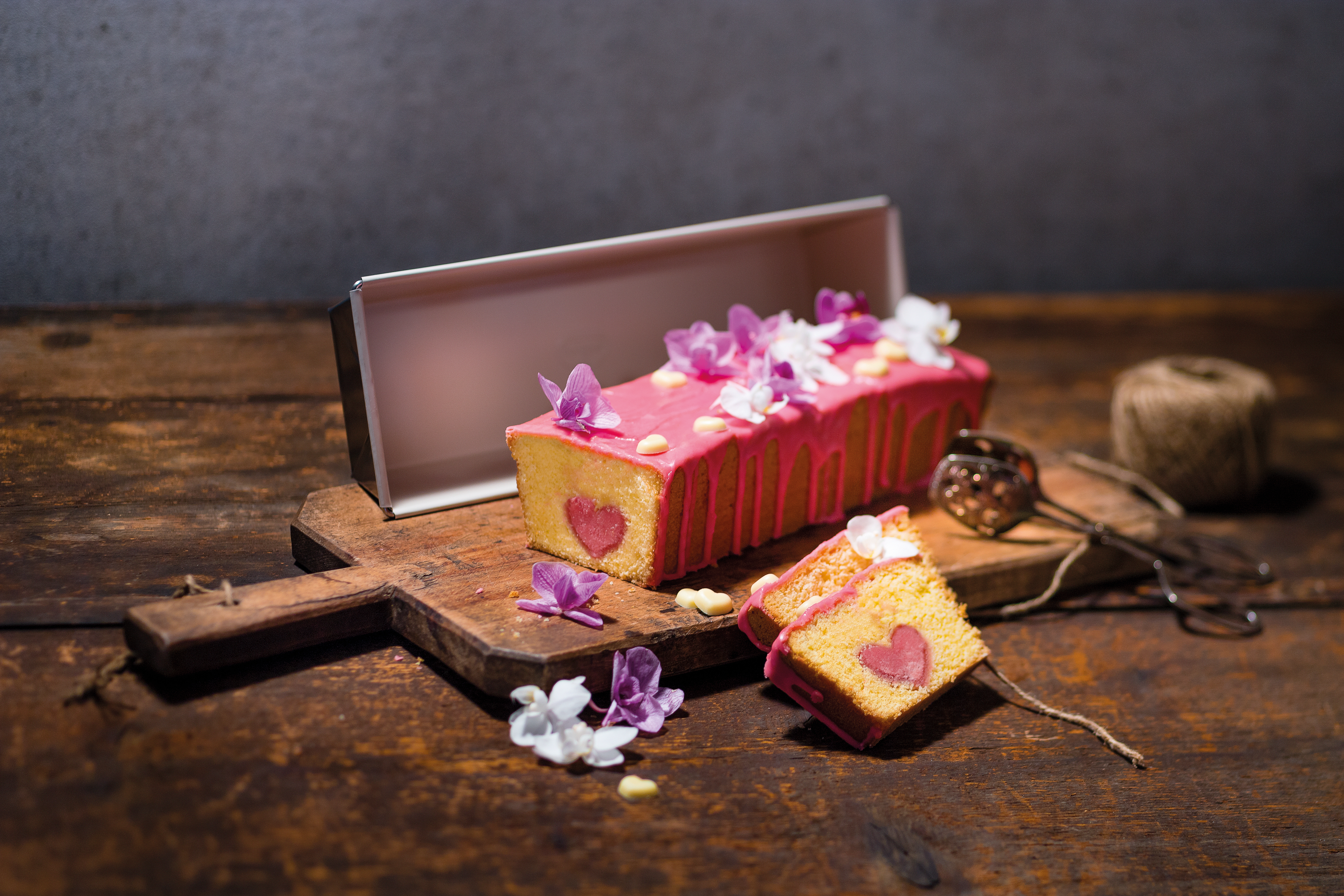 Ein rosa Kuchen mit herzförmigem Kern, garniert mit Blumen, liegt auf einem Holzbrett. Zwei Scheiben liegen davor.