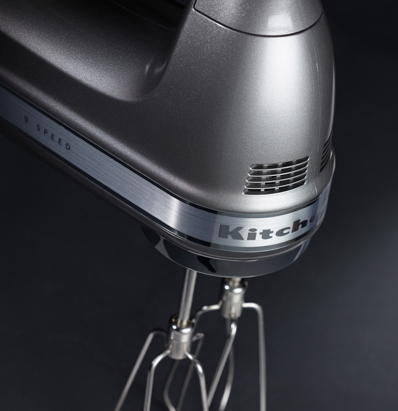 Close-up of a gray KitchenAid hand mixer with beaters, black background.