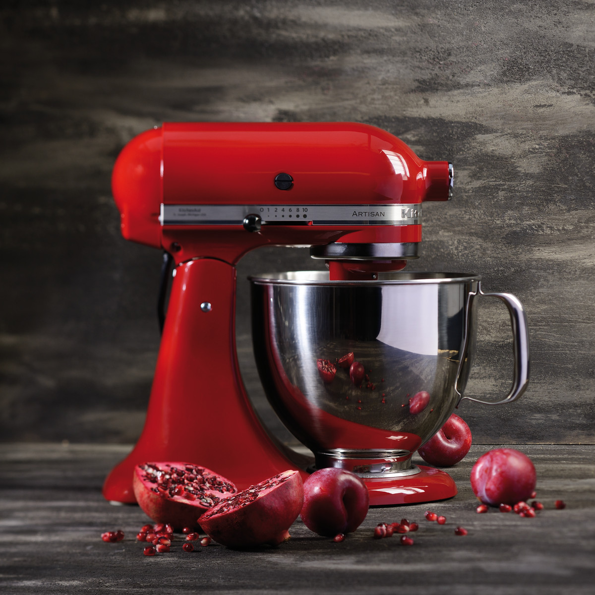 A red KitchenAid mixer with a silver bowl. Pomegranates and plums are on the wooden surface. The background is a dark wooden wall.