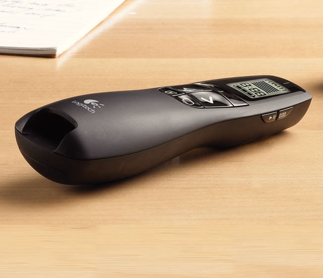 Eine schwarze Logitech-Präsentationsfernbedienung liegt auf einem Holztisch. Ein Notizblock ist teilweise im Hintergrund zu sehen.