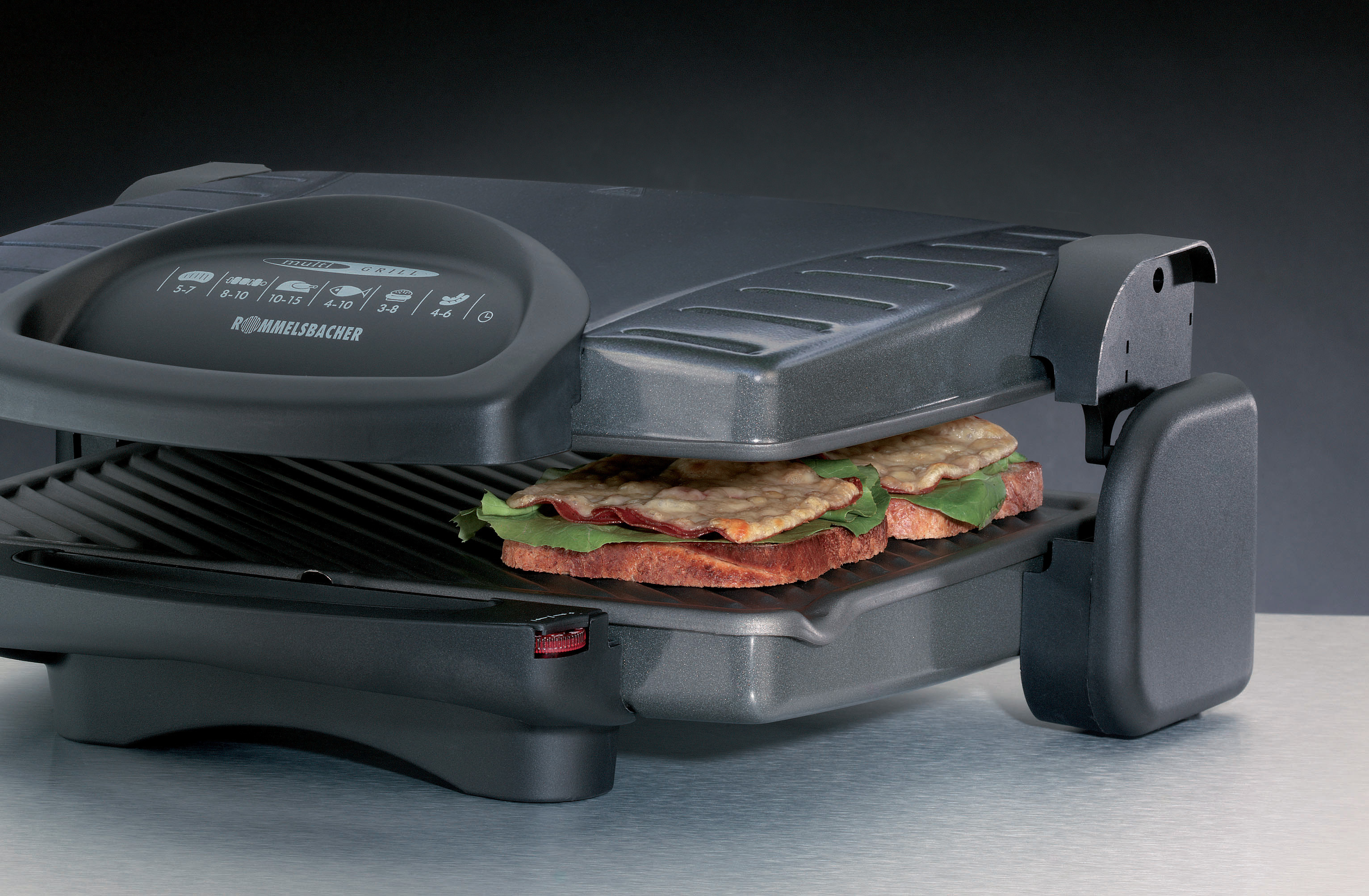 Ein geschlossener grauer Sandwichmaker mit einem Sandwich darin. Der Hintergrund ist eine graue Wand.