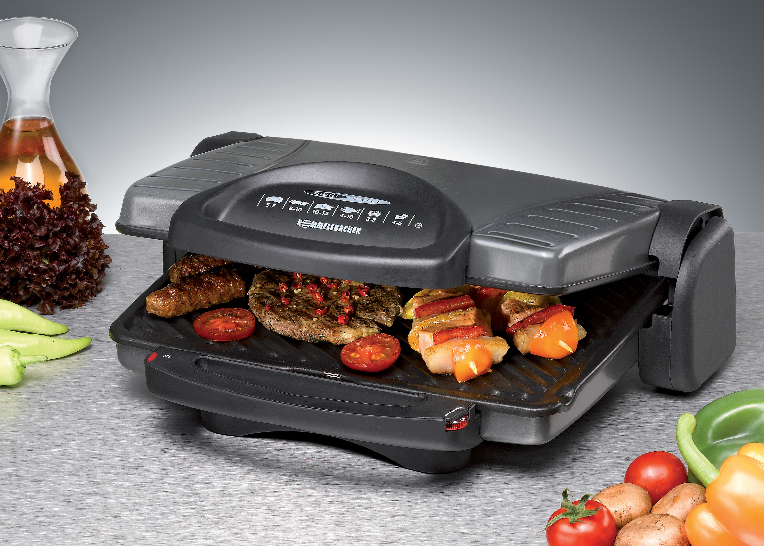 Elektrischer Grill mit darauf bratenen Speisen, darunter Würstchen, Tomaten und Spieße.