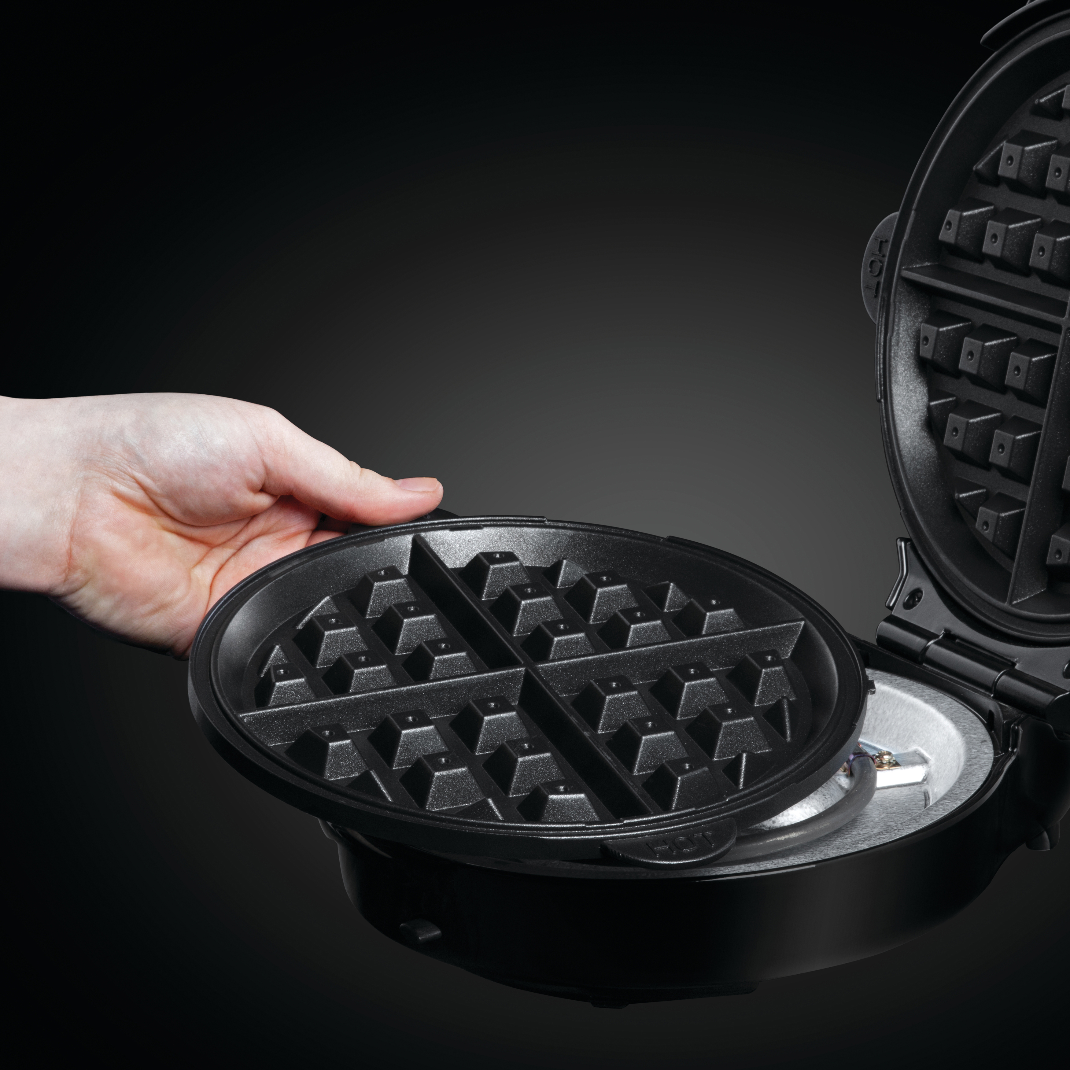 Eine Hand, die den Deckel eines Waffeleisens hält. Die Waffelform ist schwarz und geöffnet.