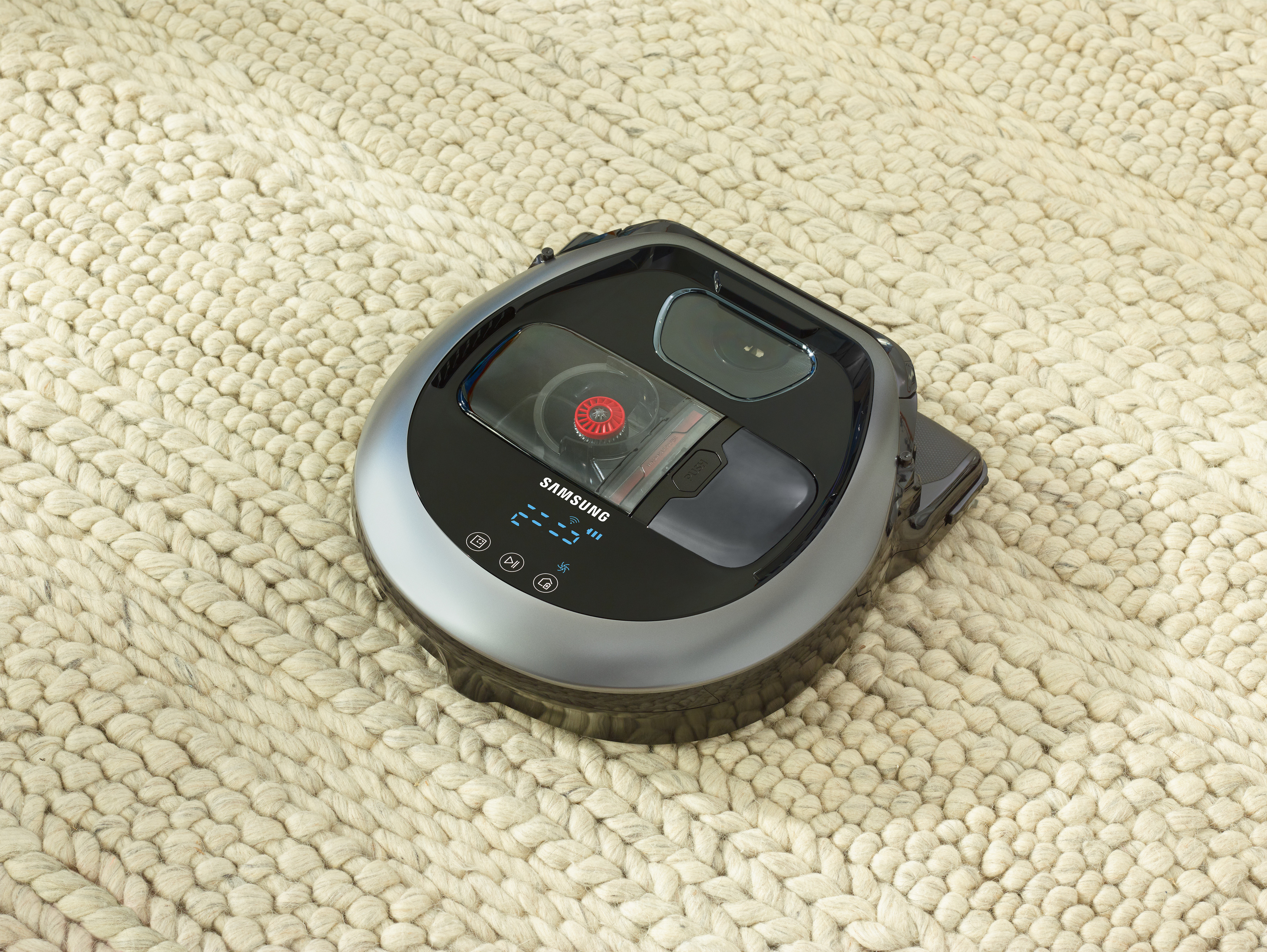 Roboter-Staubsauger auf beigem Teppich. Der Staubsauger ist schwarz und grau, mit Samsung-Logo.