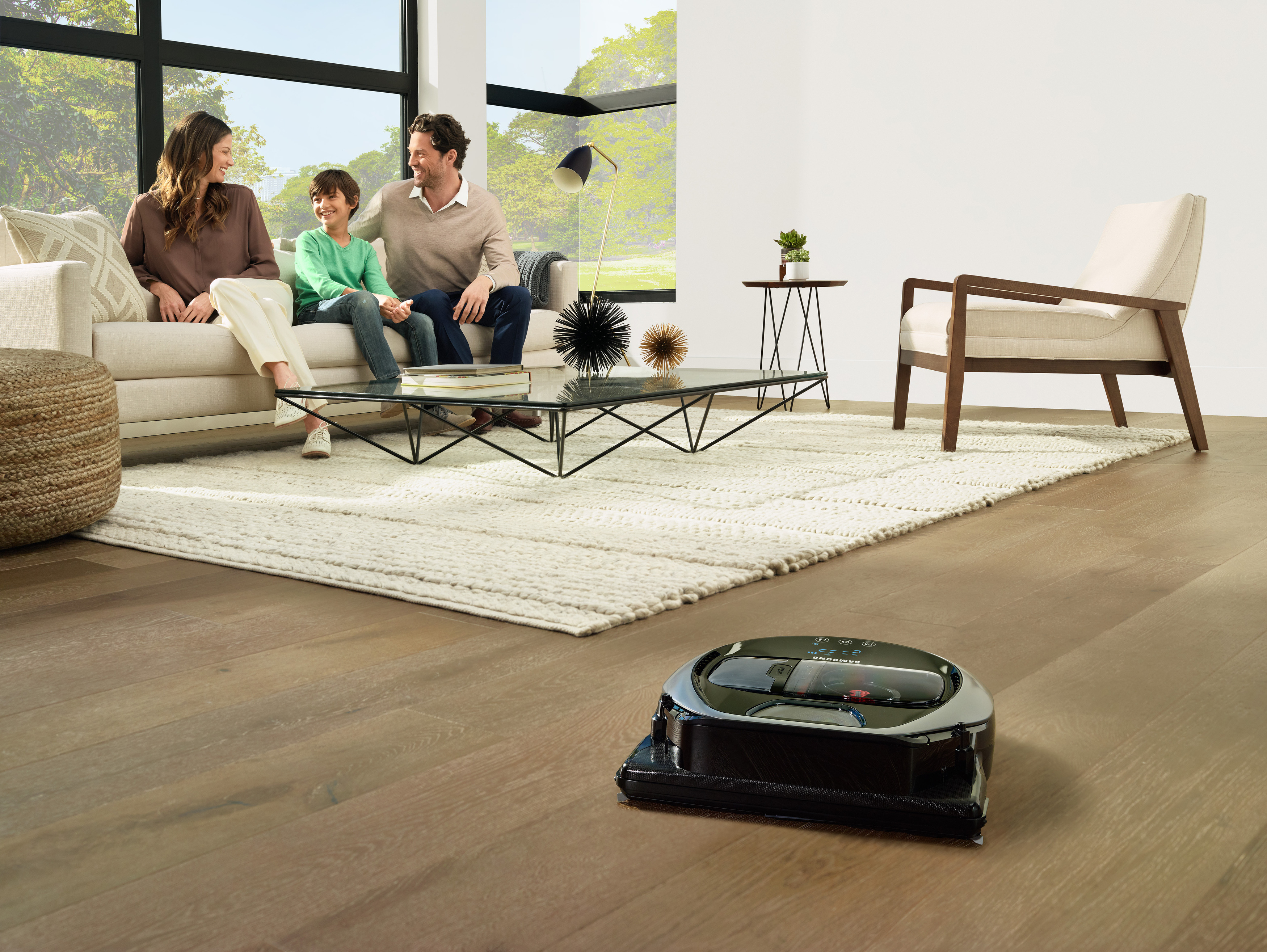 Familie auf dem Sofa, ein Roboterstaubsauger auf dem Boden, große Fenster im Hintergrund.