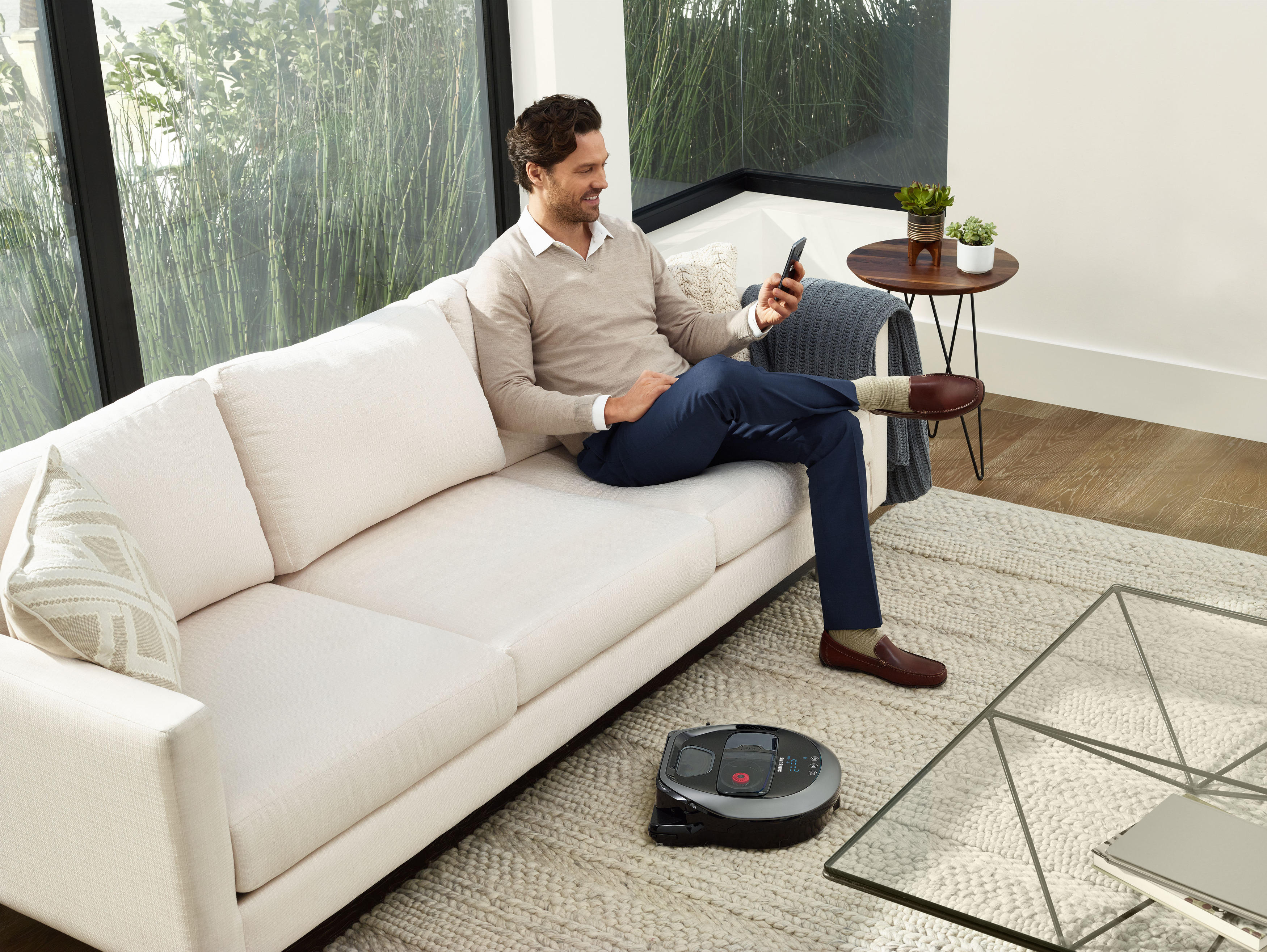 Mann in einem Wohnzimmer, der ein Telefon benutzt. Ein Roboterstaubsauger steht auf dem Boden.