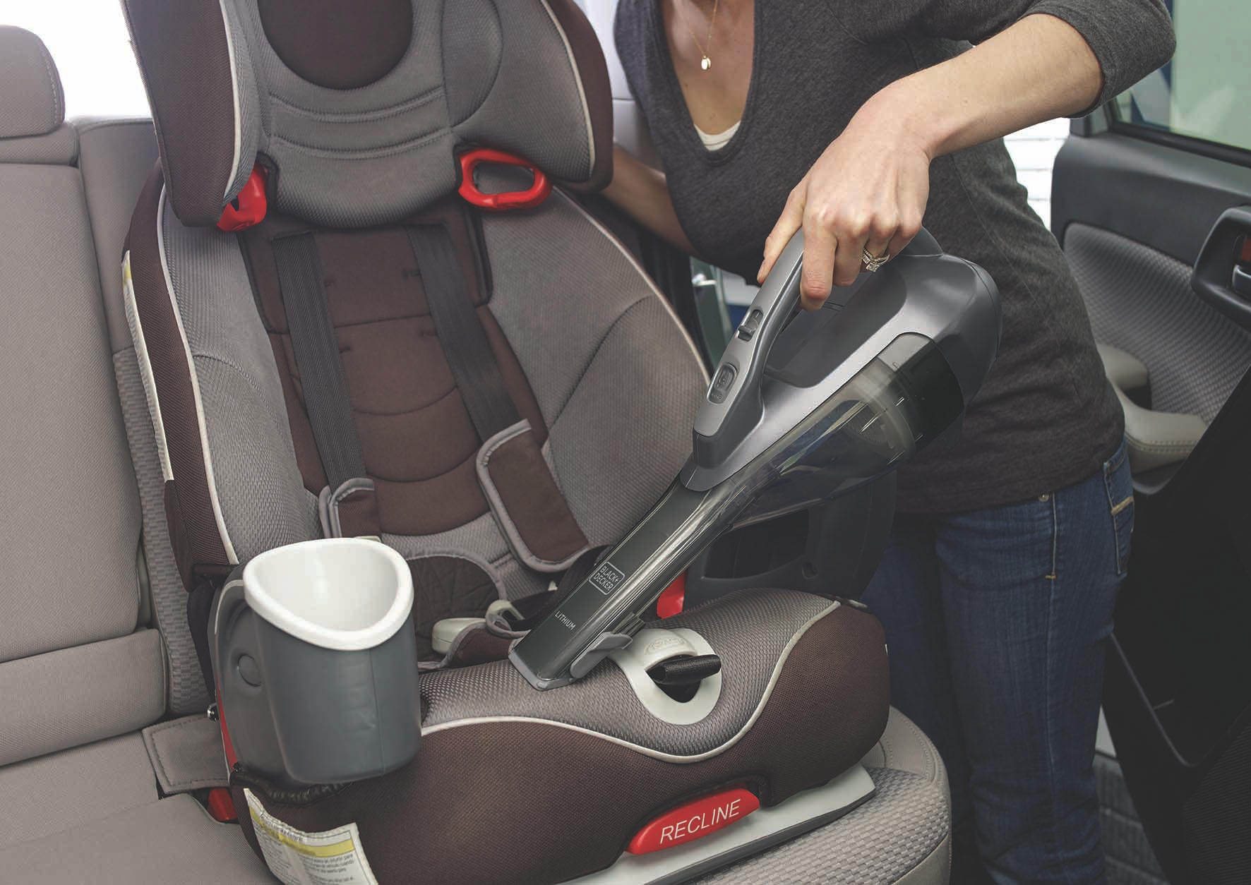 Eine Frau saugt einen Kindersitz im Auto ab. Der Sitz ist grau und braun mit roten Akzenten. Ein Becherhalter ist befestigt.