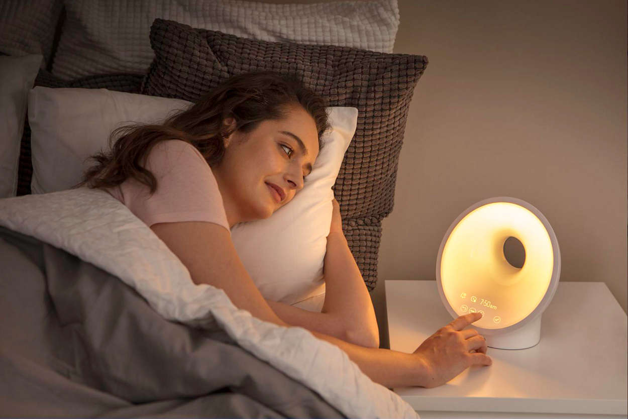 Vrouw in bed, grijpt naar een oplichtende wekker op een nachtkastje.