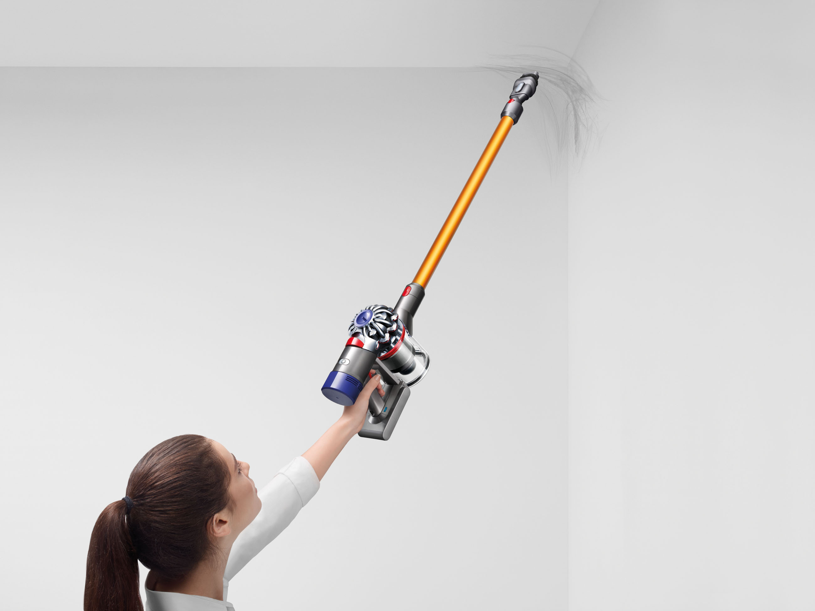 Frau benutzt orangefarbenen und silbernen Staubsauger an weißer Wand. Dunkles Haar im Pferdeschwanz. Reinigungsaktion.
