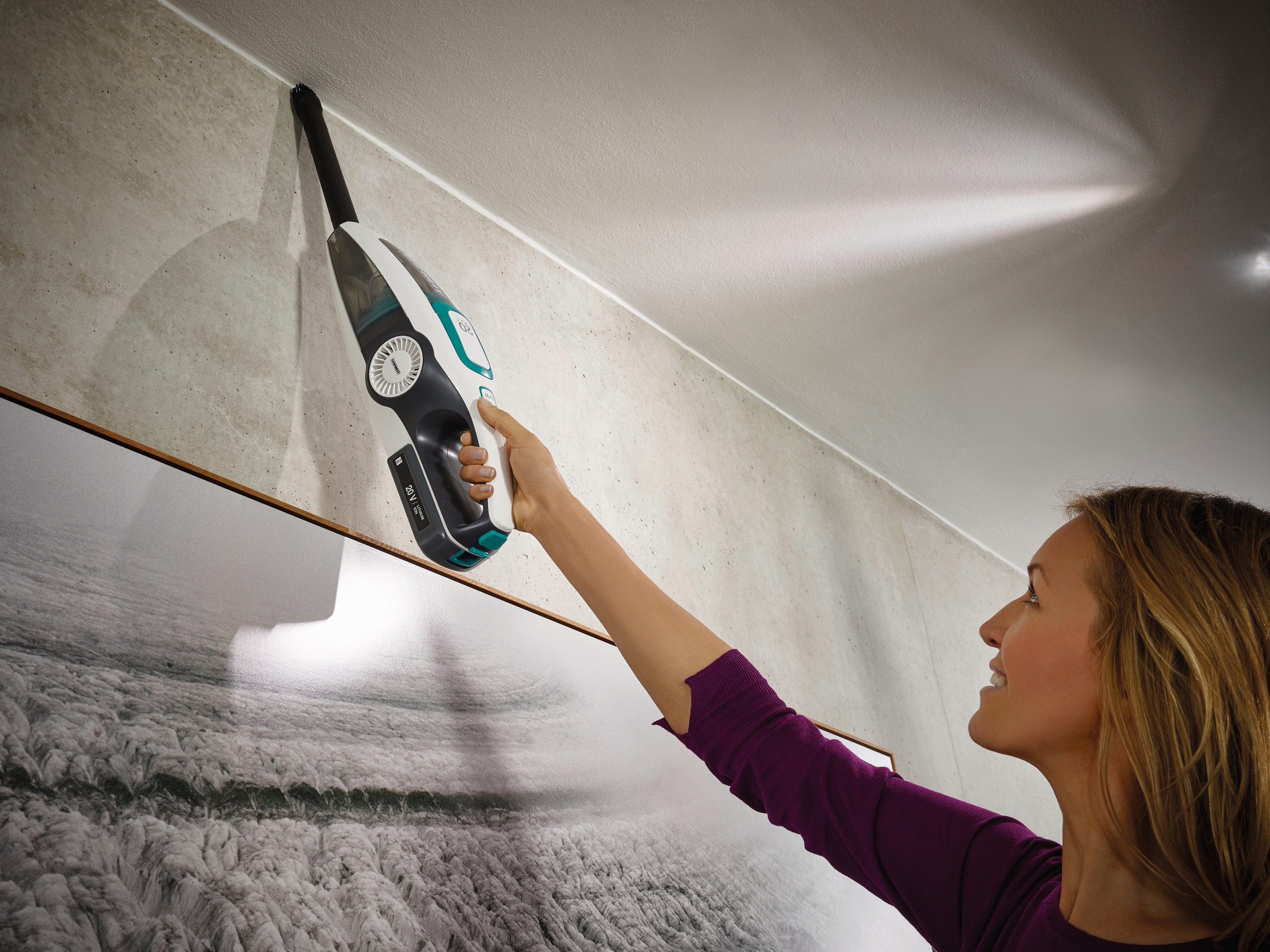 Eine Frau saugt mit einem Handstaubsauger die Wand. Sie lächelt, während sie einen weißen und tealfarbenen Staubsauger hält.