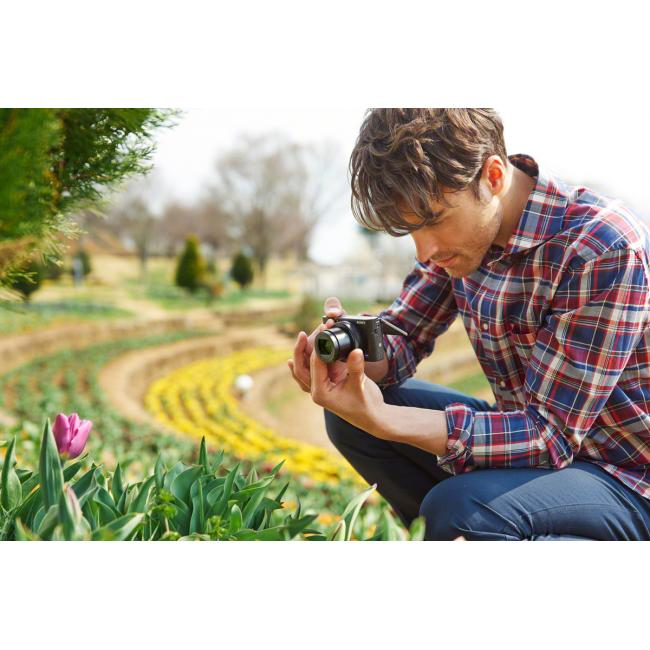 Un homme accroupi regarde un appareil photo près de fleurs, avec un champ en arrière-plan.