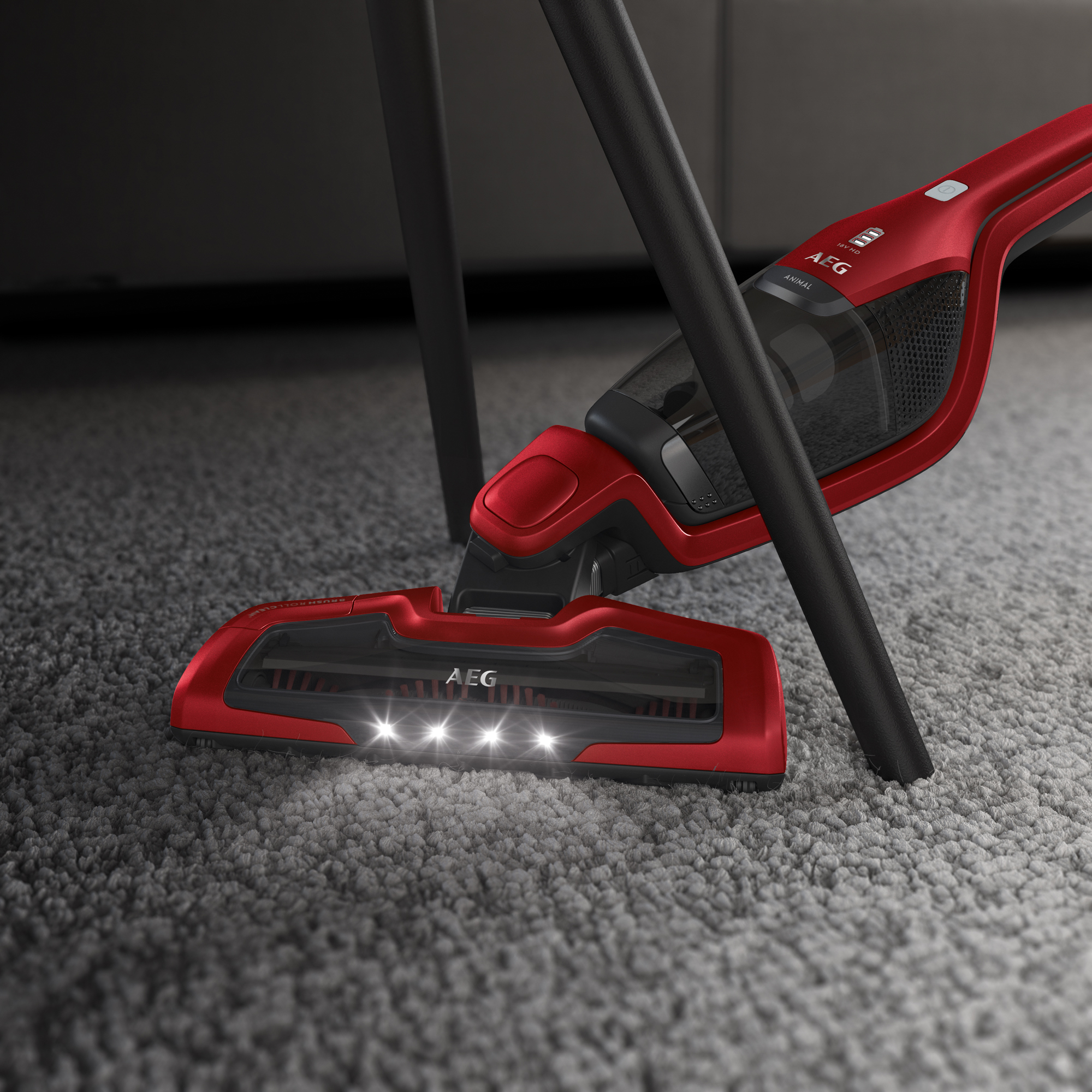 Roter AEG Animal Staubsauger reinigt einen grauen Teppich, Lichter an, schwarze Beine im Hintergrund.