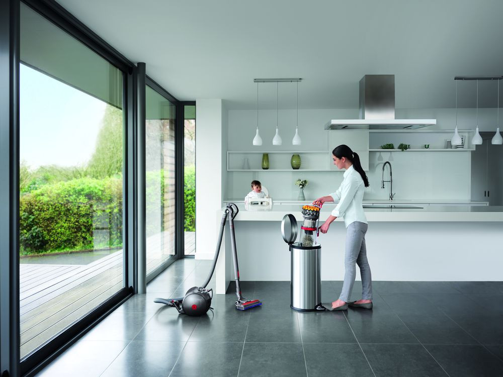 Femme utilisant un aspirateur dans une cuisine moderne, une autre personne avec un bébé en arrière-plan.