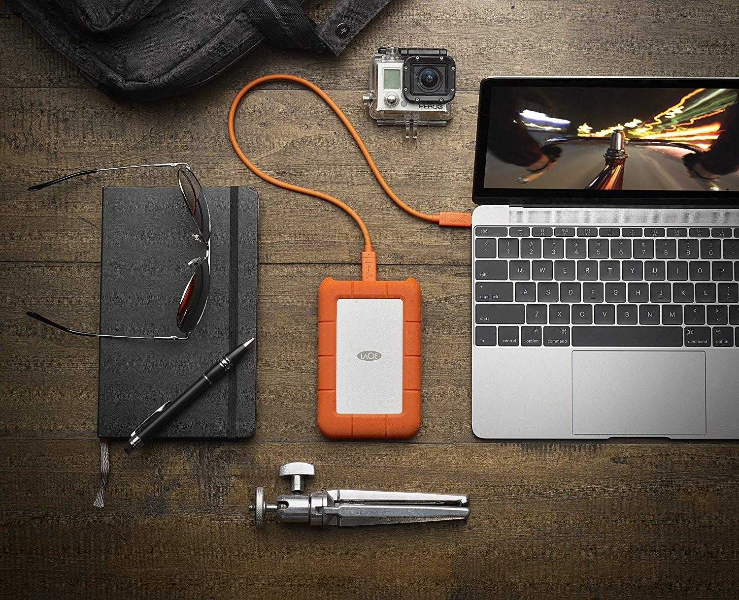 Ordinateur portable, appareil photo, disque dur externe, lunettes de soleil, stylo et trépied sur une table en bois.