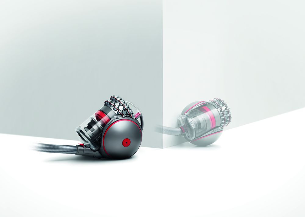 A gray and red Dyson vacuum cleaner stands on a white surface, with its reflection visible. 
