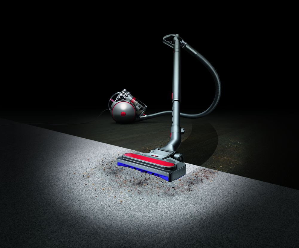 A Dyson vacuum cleaner on a carpeted floor. The vacuum is black and red, with a dark background.