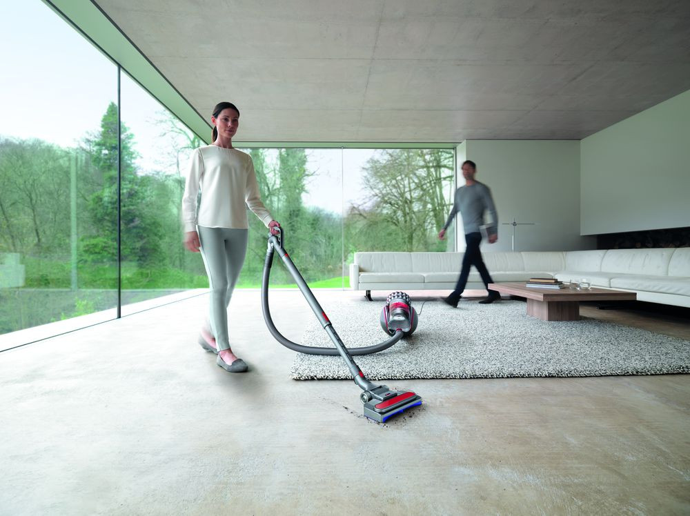 Woman vacuums a room. A man walks past. A Dyson vacuum is on the floor.